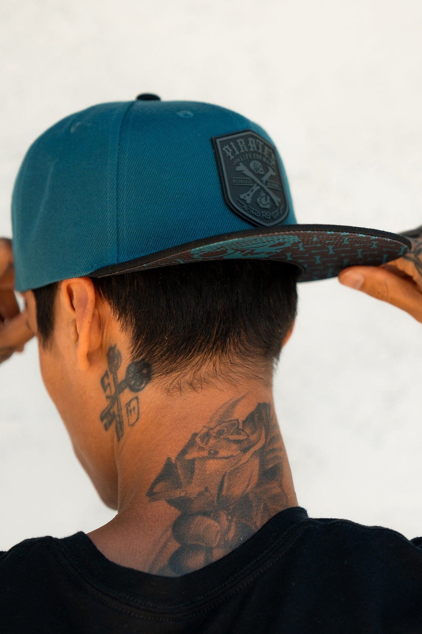 A tattooed individual in a black shirt adjusts their Pirates Life Teal Snapback by Relentless Betrayal backward, standing against a neutral wall, exuding the pirate lifestyle.