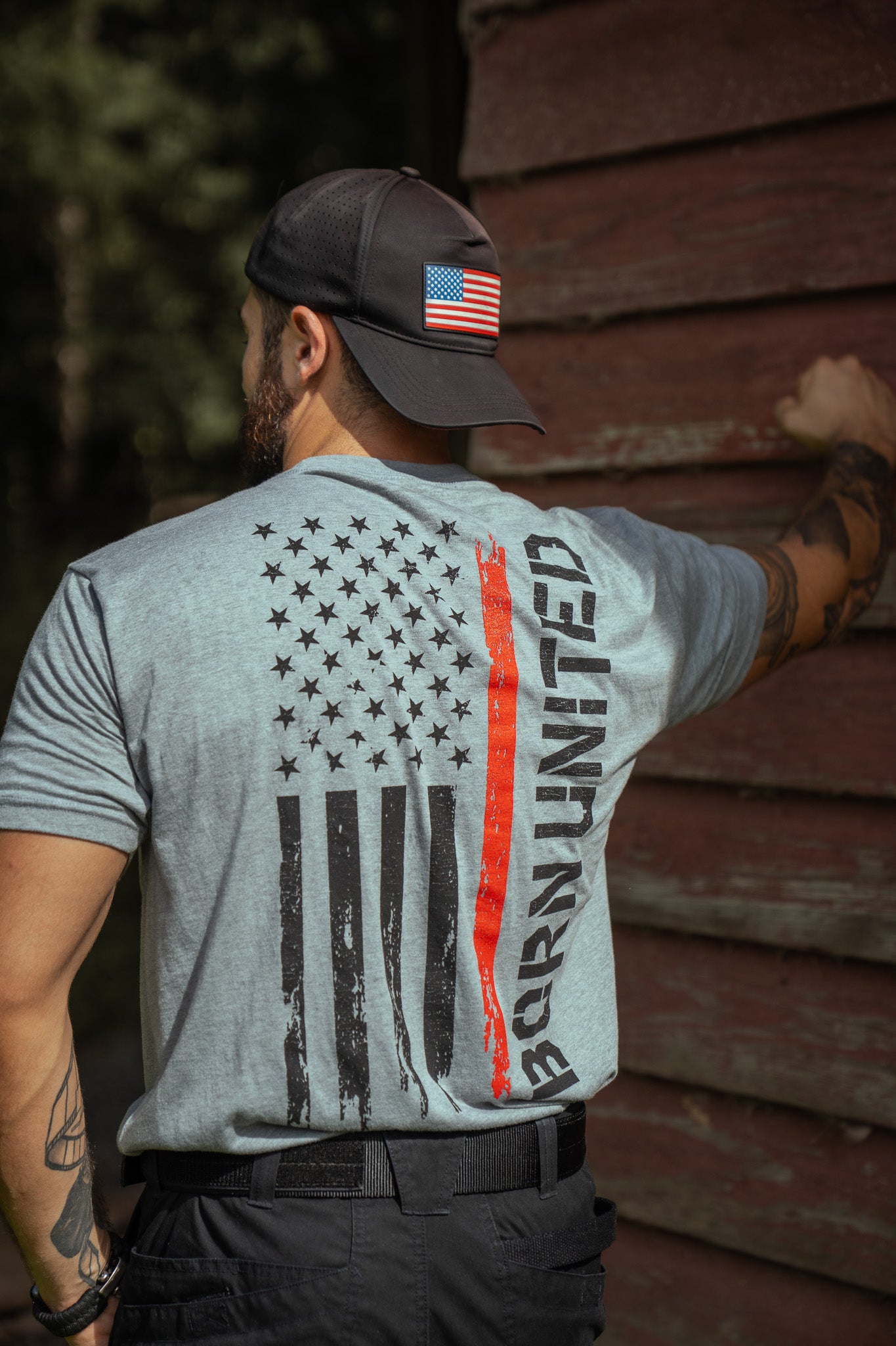 A man in a backward cap with a U.S. flag patch stands by a wooden wall, wearing the Born United Private Label Red Line Flag Tee featuring a distressed U.S. flag with a red stripe and BRAVERY printed beside it.