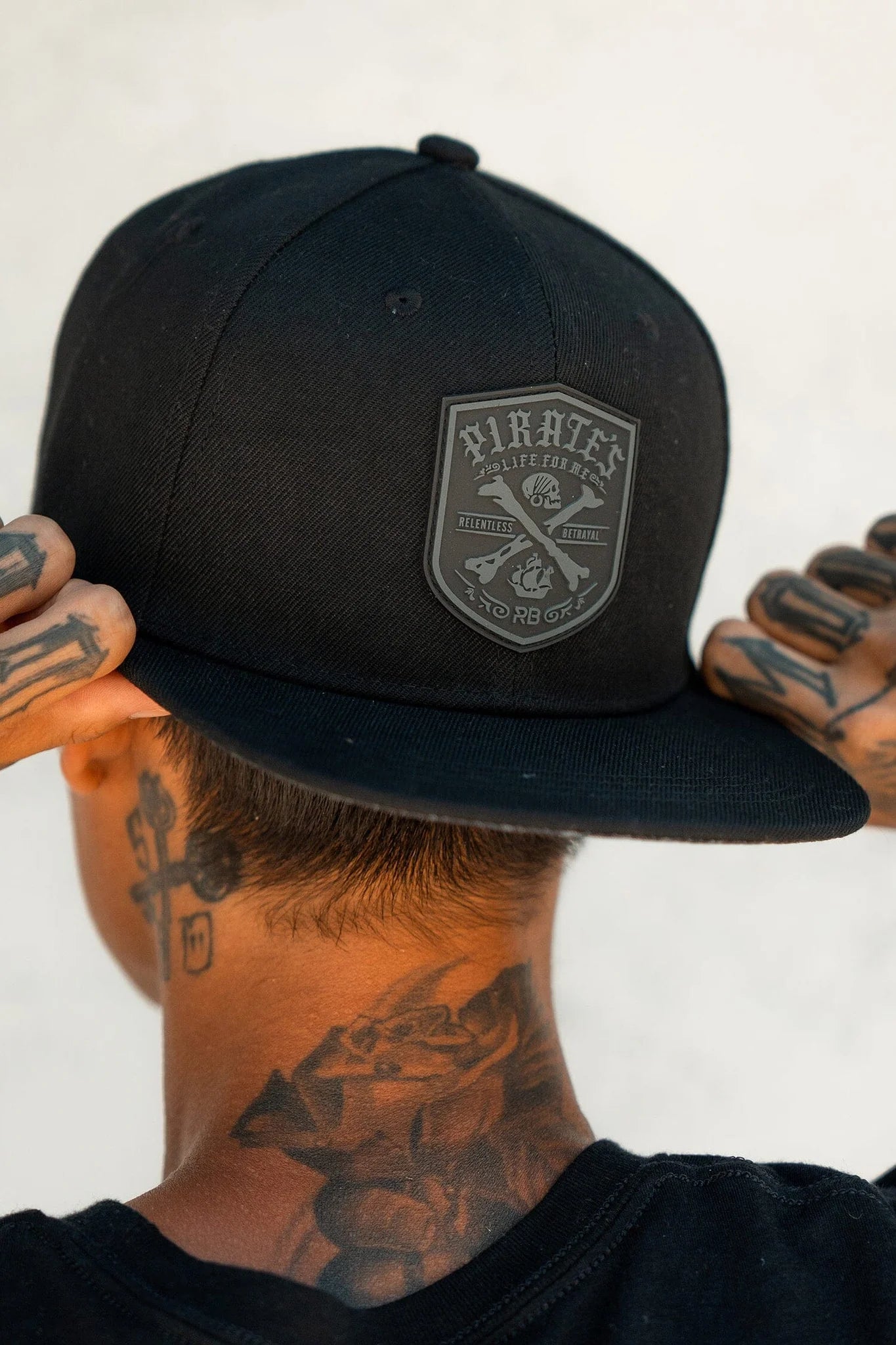 A person wears the Pirates Life Black Snapback by Relentless Betrayal, featuring a custom PVC rubber patch, viewed from behind. Tattoos are visible on their neck and hands against a plain, light-colored wall background.