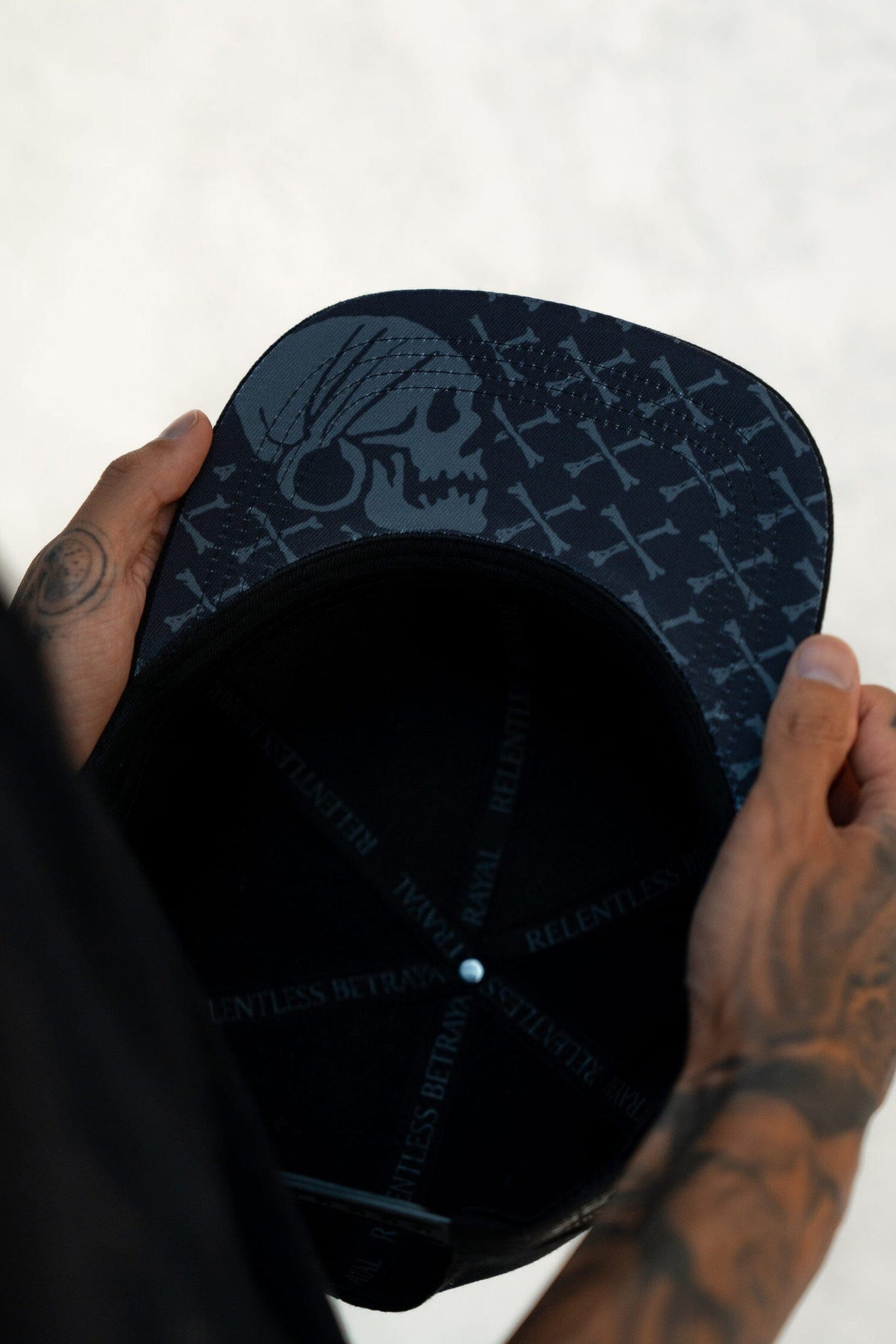 Tattooed hands hold the Pirates Life Black Snapback by Relentless Betrayal. This custom PVC rubber patch hat includes a gray skull design with cross patterns under its black cap, and RELENTLESS on the sweatband. It features an adjustable snapback for comfort.