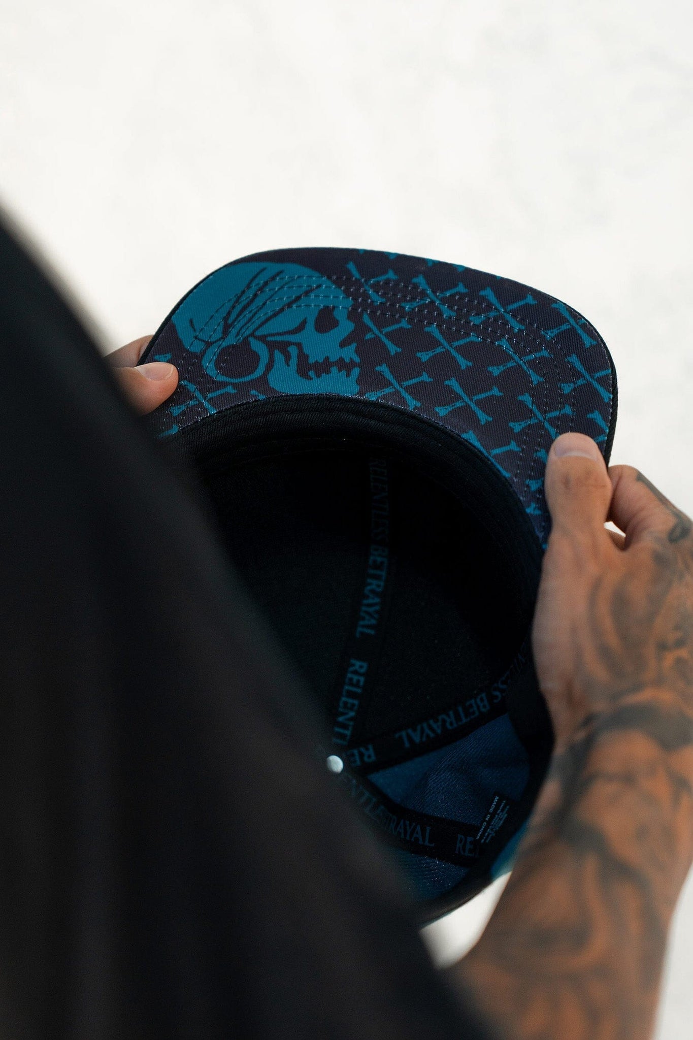 A tattooed individual holds a Pirates Life Teal Snapback by Relentless Betrayal, featuring a skull design under the brim and Rebels Betrayal printed inside. The adjustable snapback complements the hint of a pirate lifestyle on a plain light background.