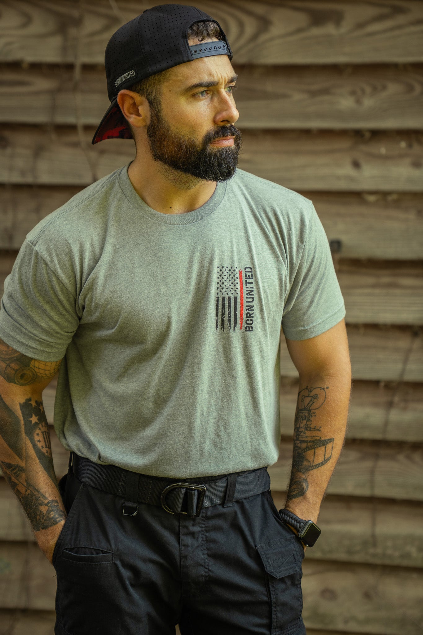 A bearded, tattooed man in a gray Red Line Flag Tee by Born United Private Label and a backwards black cap stands outdoors by a wooden wall, looking to the side.