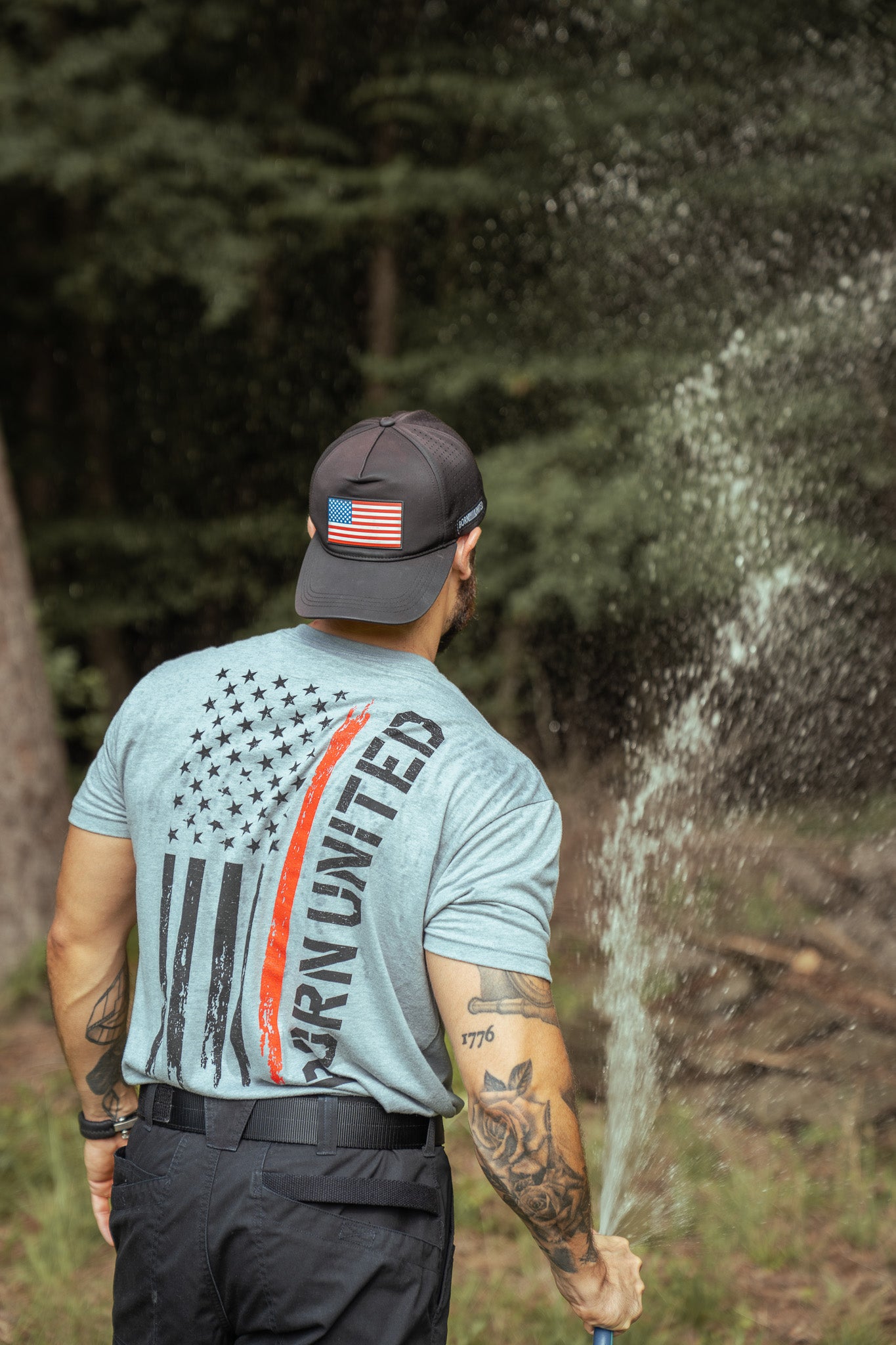 A tattooed man, in a cap with an American flag patch and a Born United Private Label Red Line Flag Tee, sprays water outdoors near trees.