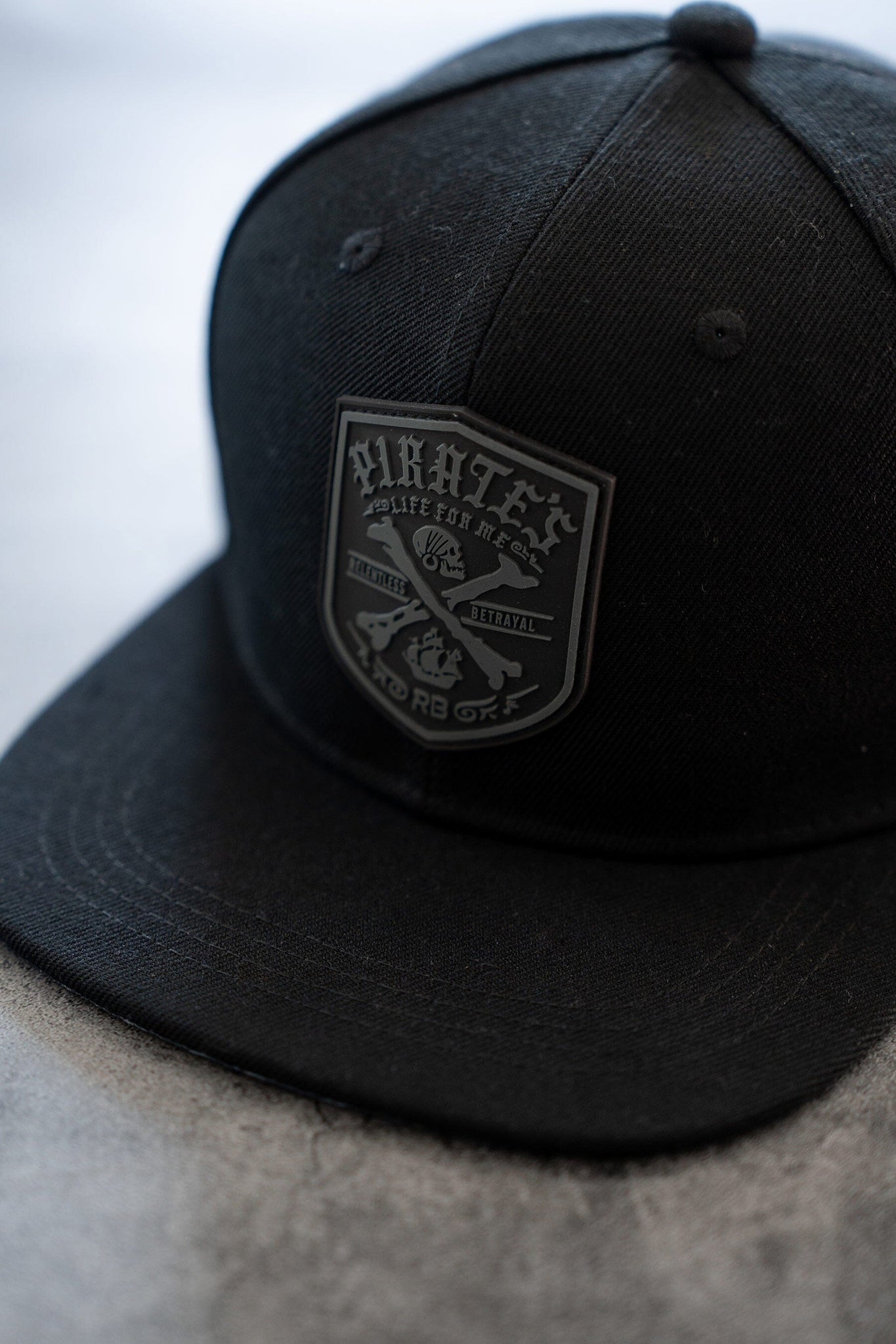 The Pirates Life Black Snapback by Relentless Betrayal features a custom PVC rubber patch with decorative text, a weapon symbol, and Hope or Die. It has a snapback enclosure and is artfully placed on a textured surface.