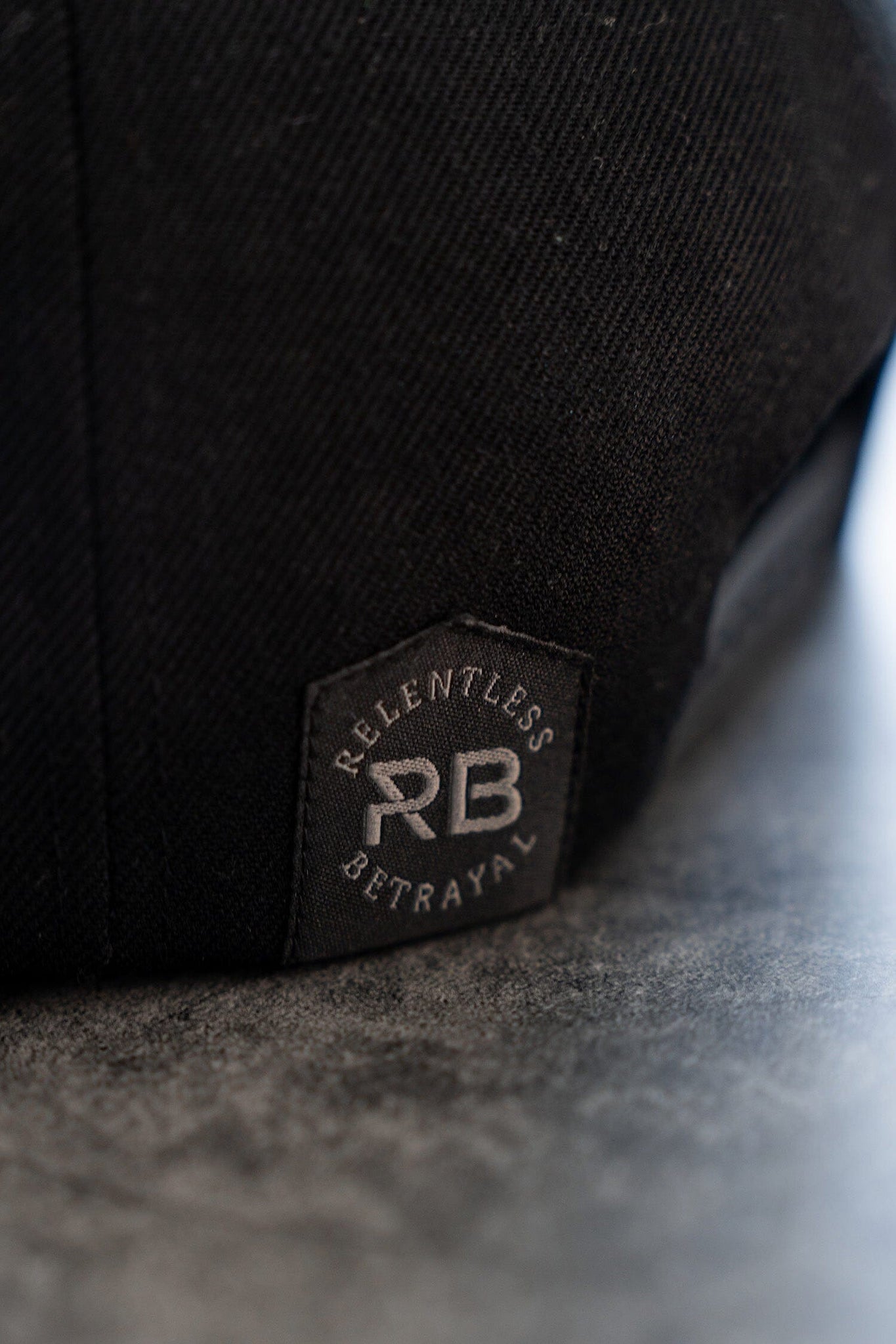 Close-up of the Pirates Life Black Snapback by Relentless Betrayal featuring Relentless Betrayal and RB on a custom PVC patch. The snapback closure ensures a snug fit, resting on a smooth, dark gray surface.