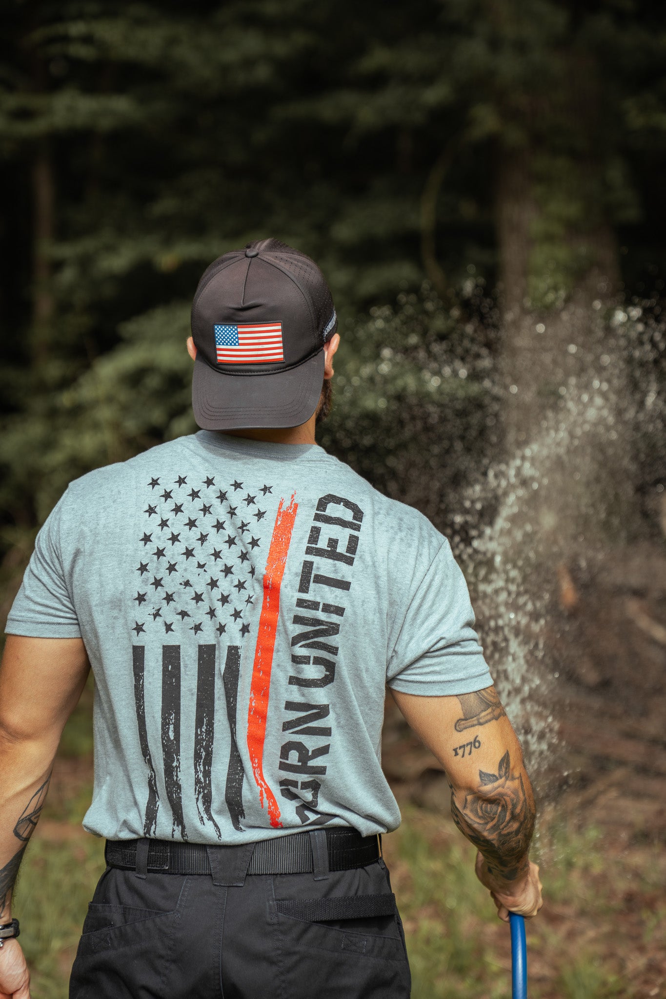A man wearing a cap with an American flag patch and a Born United Private Label Red Line Flag Tee, featuring a black-and-white flag and “UNITED,” waters outdoor plants, standing among trees, seen from behind.