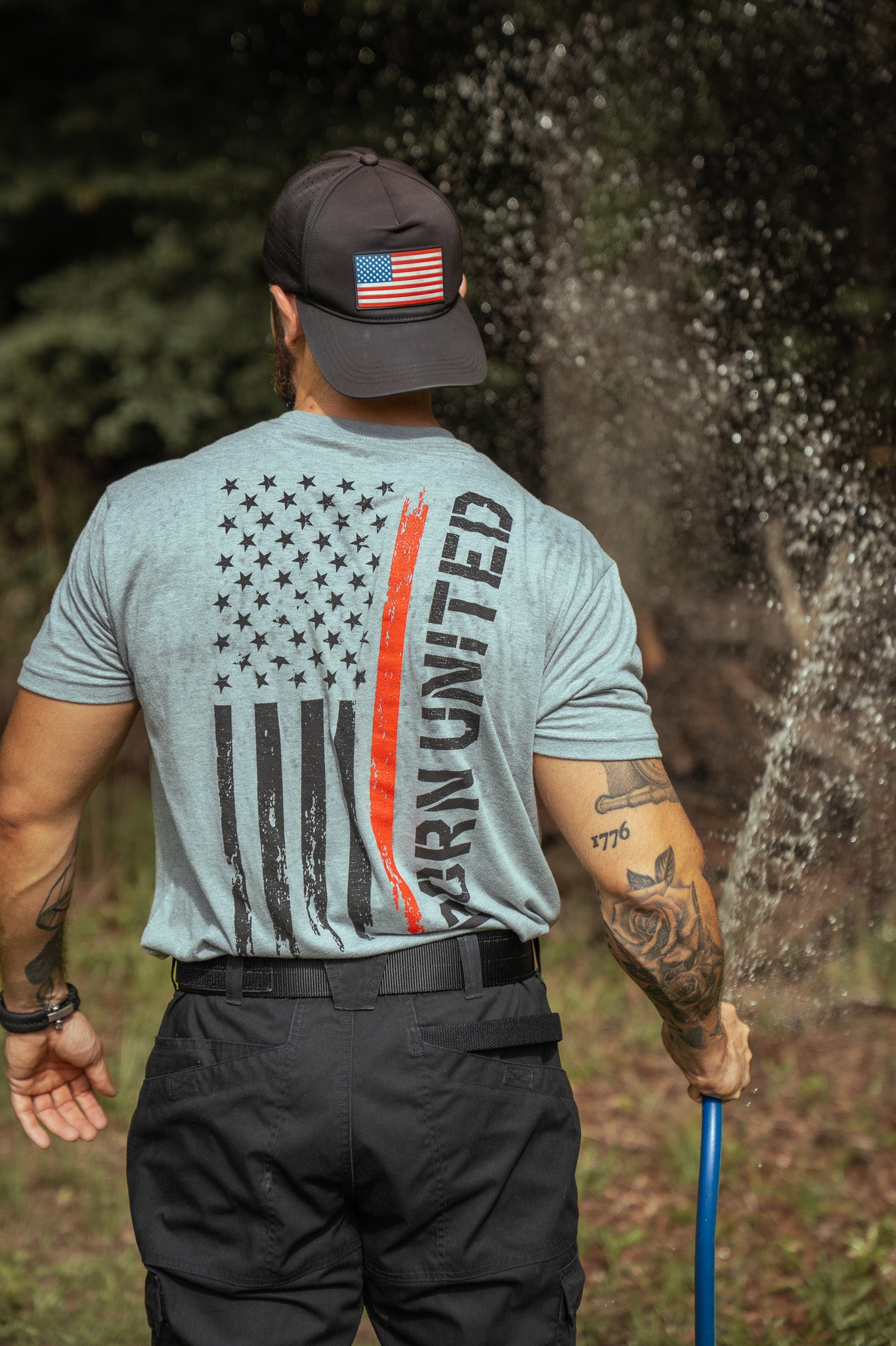 A man seen from behind wears a Born United Private Label Red Line Flag Tee featuring a stylized American flag and UNITE in bold letters, while watering grass outdoors with a blue hose amidst trees and greenery.