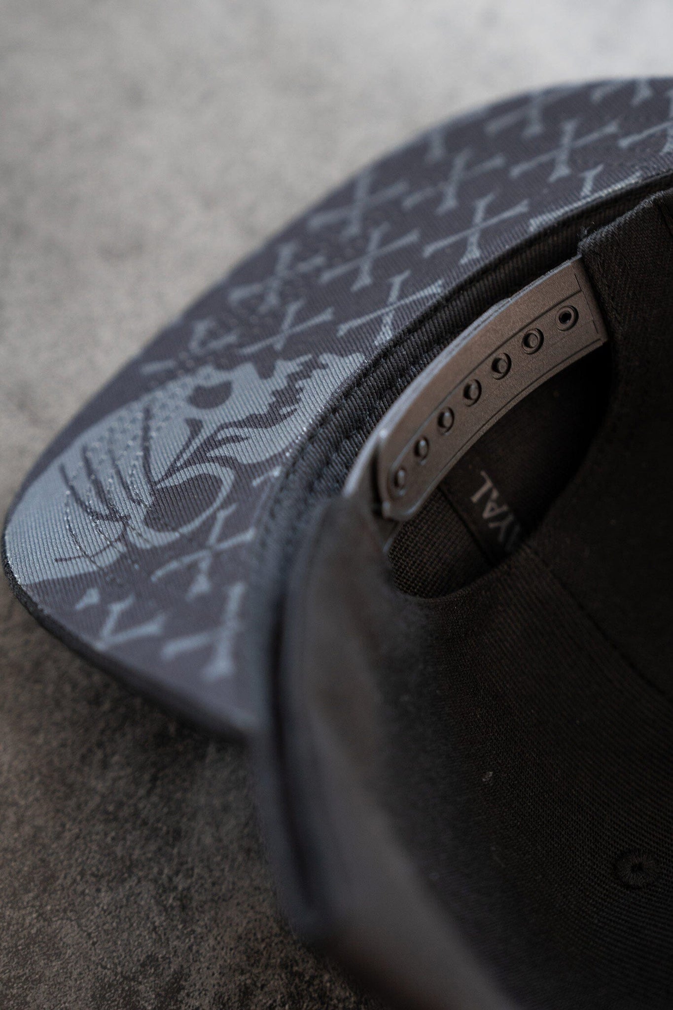 Close-up of the Pirates Life Black Snapback cap by Relentless Betrayal on a gray surface. The inner brim has abstract shapes and letters, while an adjustable strap with fastener holes highlights the snapback closure. Part of FINAL is visible inside.