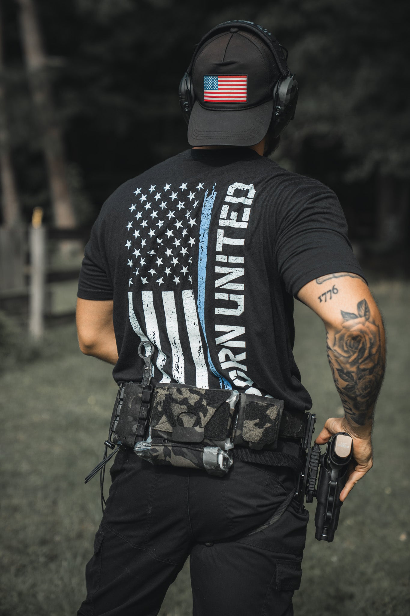 Wearing a Born United Private Label Blue Line Flag Tee and a black cap, a tattooed person with a utility belt stands outdoors holding a handgun, proudly showing support for law enforcement.