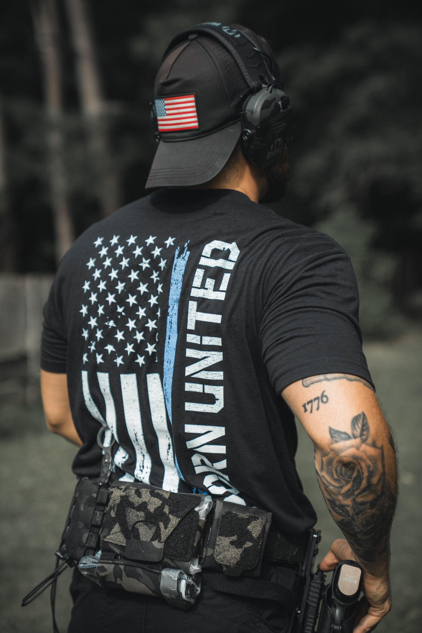 A person outdoors wears a black cap and the Born United Private Label Blue Line Flag Tee, featuring a blue line American flag and Born United text. They carry tactical gear, have a holstered weapon, and display left arm tattoos, showing Back the Blue support.