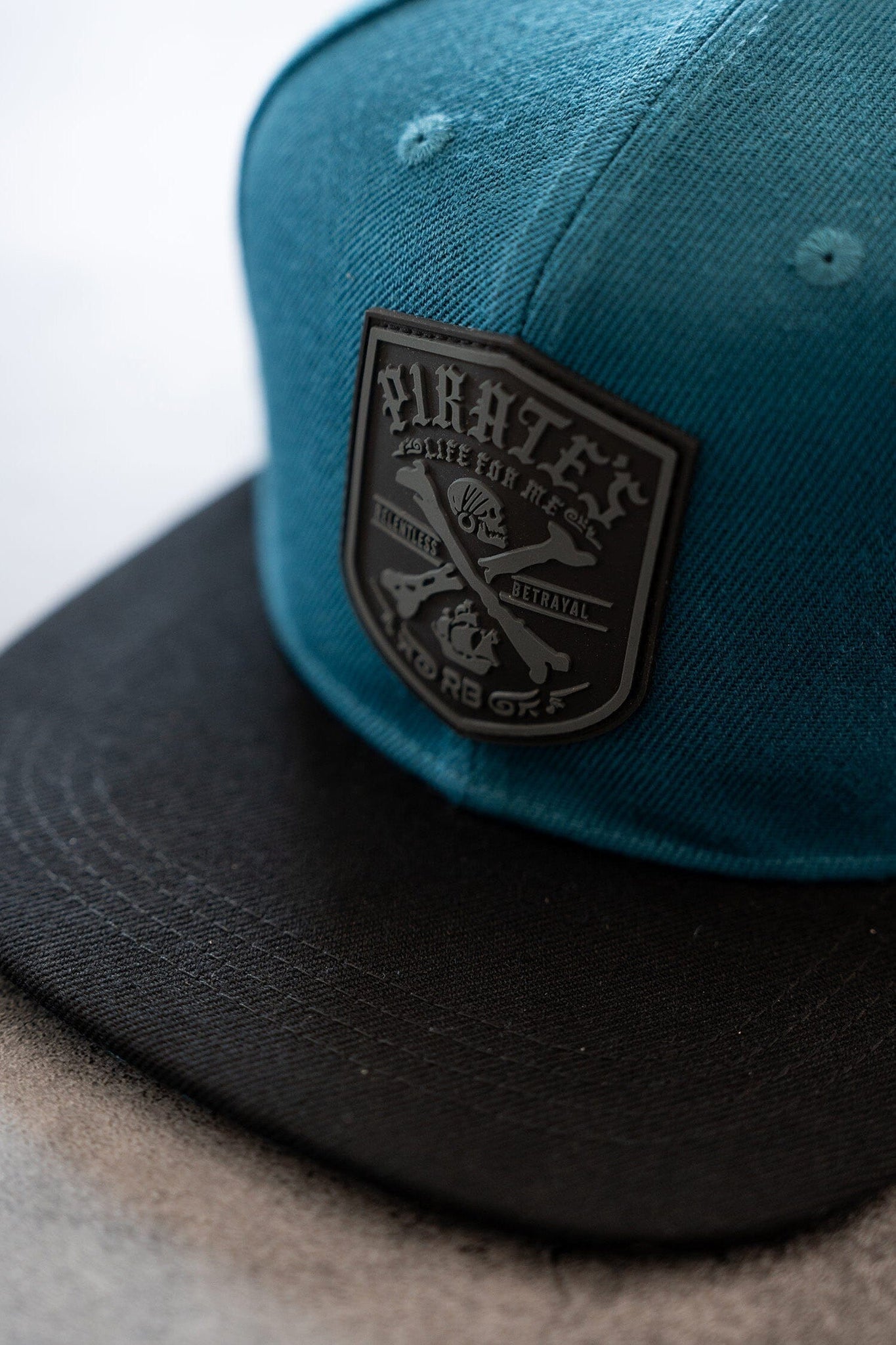 A close-up of the Pirates Life Teal Snapback by Relentless Betrayal features a teal and black design with a PVC rubber patch. The patch, detailed with crossed elements and text, evokes a vintage pirate lifestyle. The snapback is on a light-colored surface.