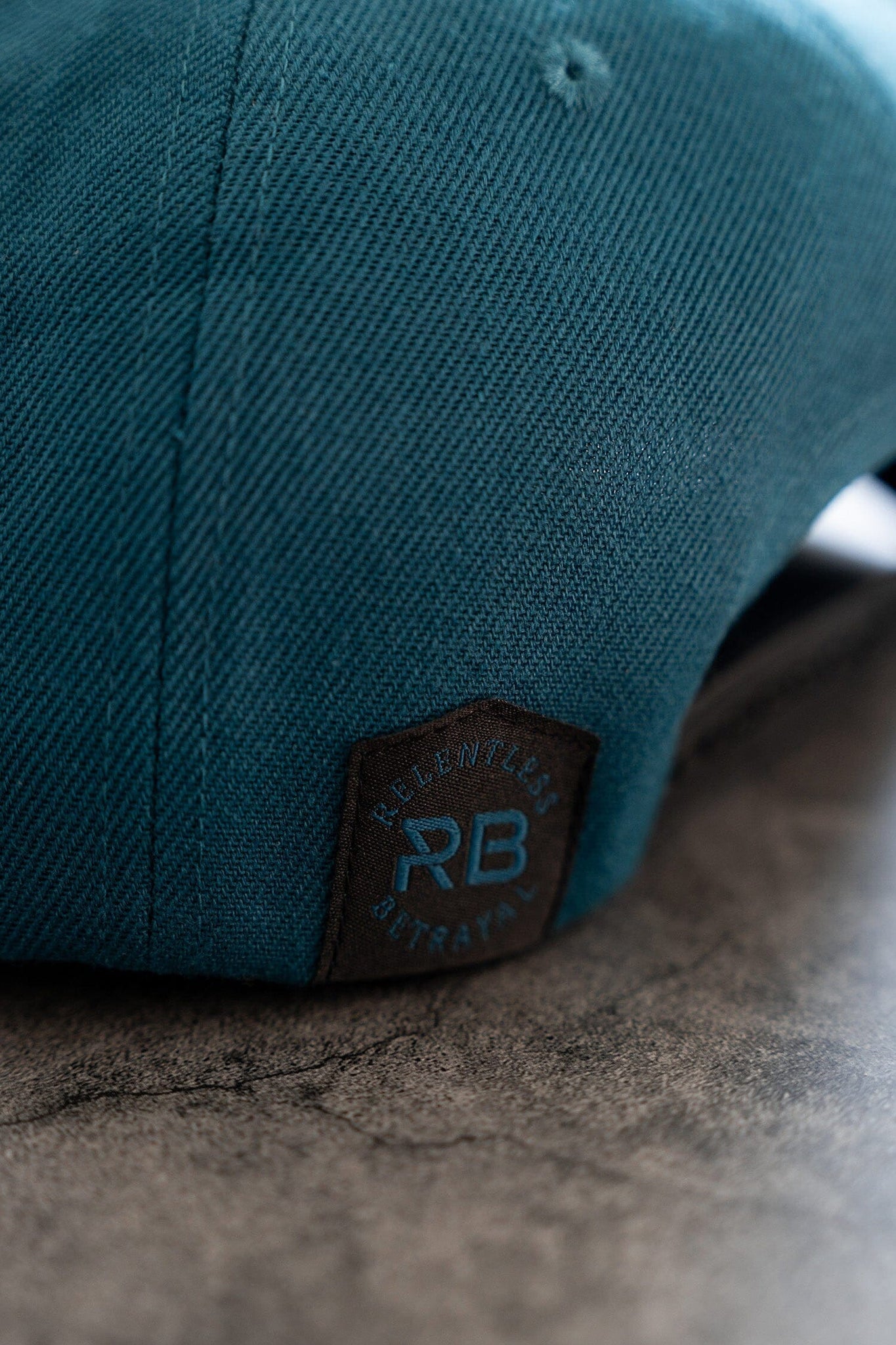 Close-up of the Pirates Life Teal Snapback by Relentless Betrayal, featuring a black tag with RB and RELENTLESS BETRAYAL in blue stitching. The cap embraces a pirate lifestyle and rests on a textured gray surface.
