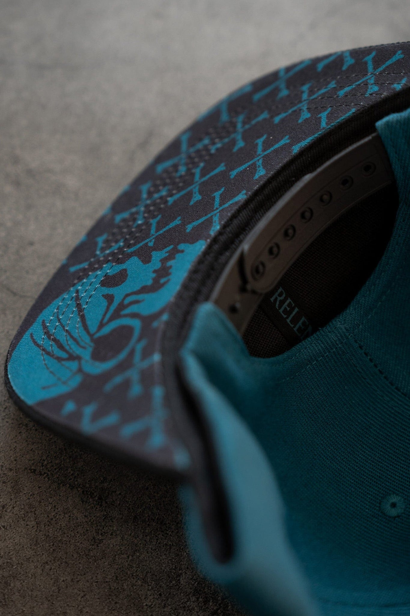 Close-up of the underside of the Pirates Life Teal Snapback by Relentless Betrayal, featuring a snapback closure. The brim showcases a blue abstract claw design with geometric patterns, while the interior reveals its structure and fabric against a textured gray background.