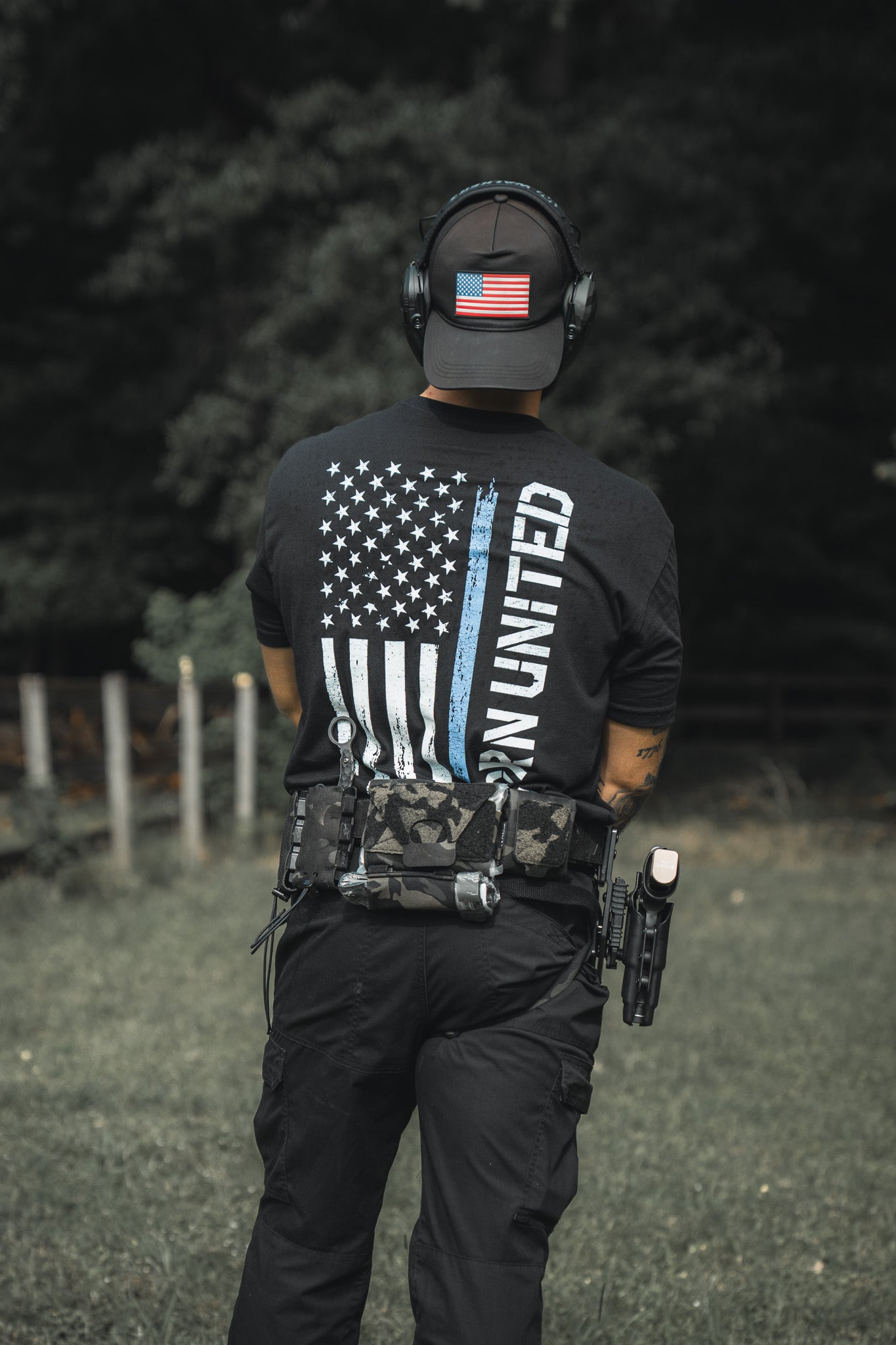 Outdoors, a person wears black tactical gear with a Born United Private Label Blue Line Flag Tee featuring a blue-striped American flag and “UNITED” text, plus an American flag patch cap, ear protection, and a holstered handgun.