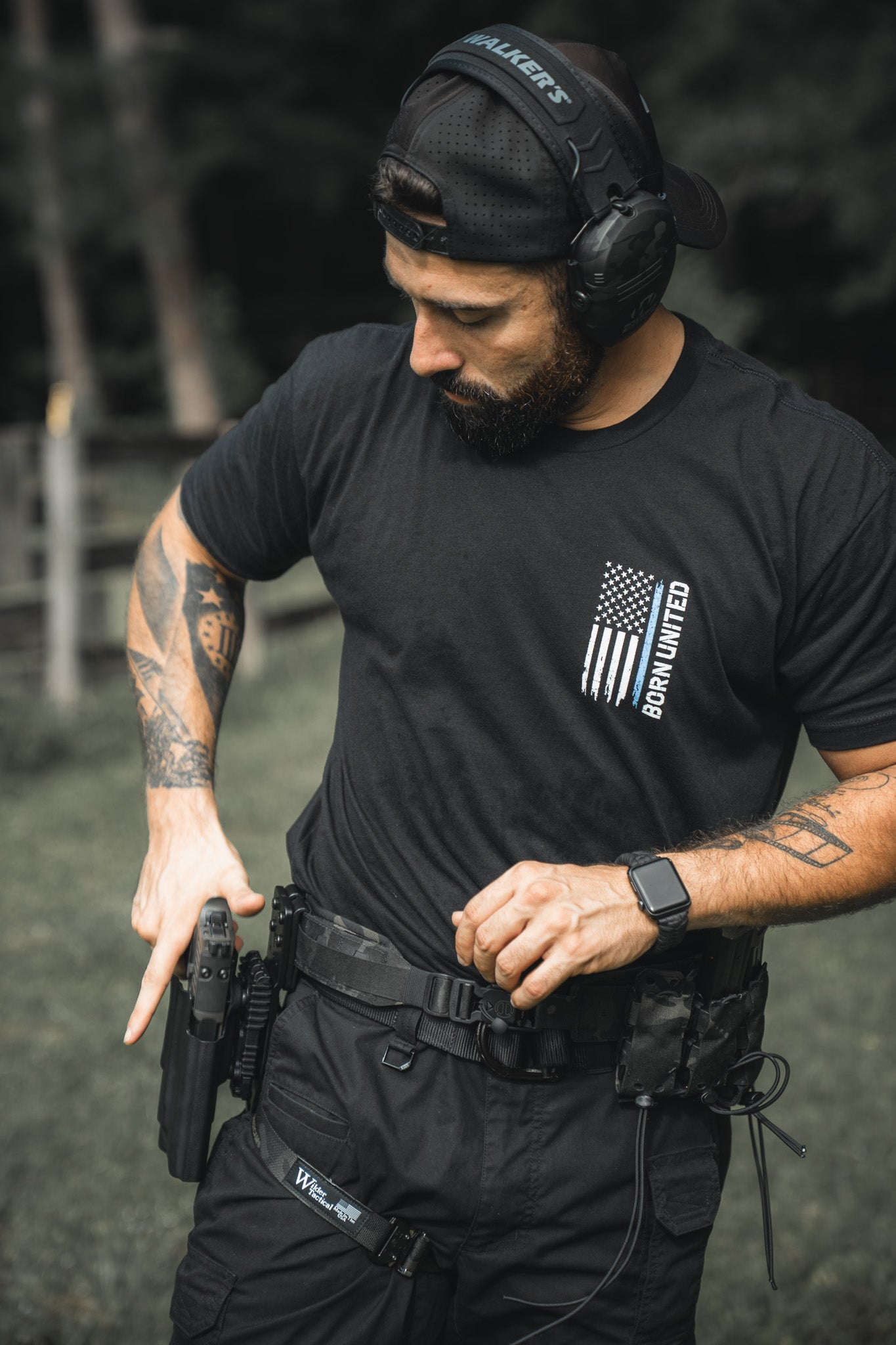 A man in a Born United Private Label Blue Line Flag Tee, cap, headphones, and tactical gear practices drawing his handgun outdoors, displaying strong police support.