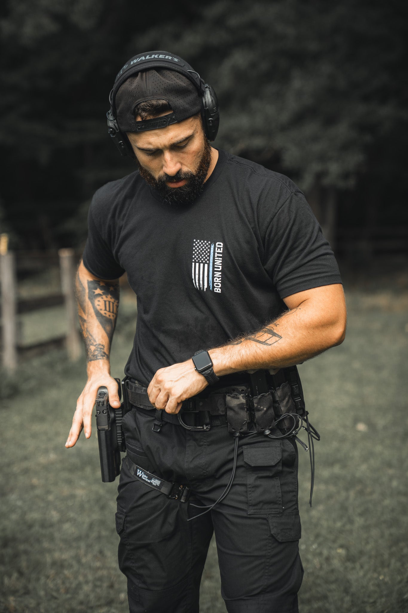 A bearded man in a Born United Private Label Blue Line Flag Tee, black cap worn backwards, black cargo pants, and tactical gear stands outdoors, adjusting his belt with one hand and holding a handgun in the other.