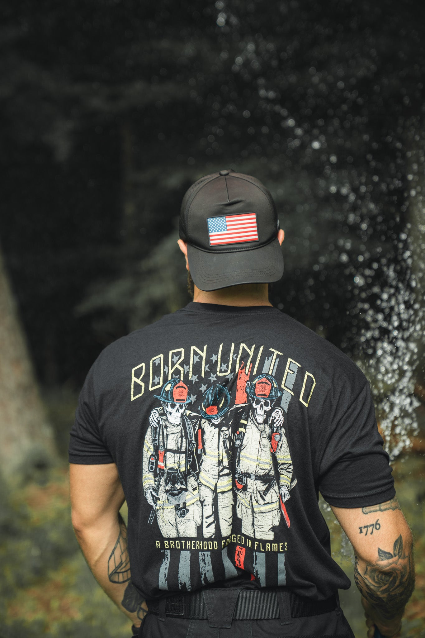 A person outdoors wears a Born United Private Label Brotherhood Forged In Flames black T-shirt with three firefighters and a black cap featuring a U.S. flag patch.