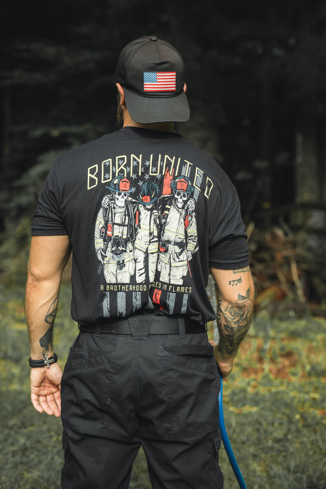 A man wearing a Born United Private Label Brotherhood Forged In Flames t-shirt and a black cap with an American flag stands outdoors, facing away. Tattoos cover his arms as he holds a blue hose.