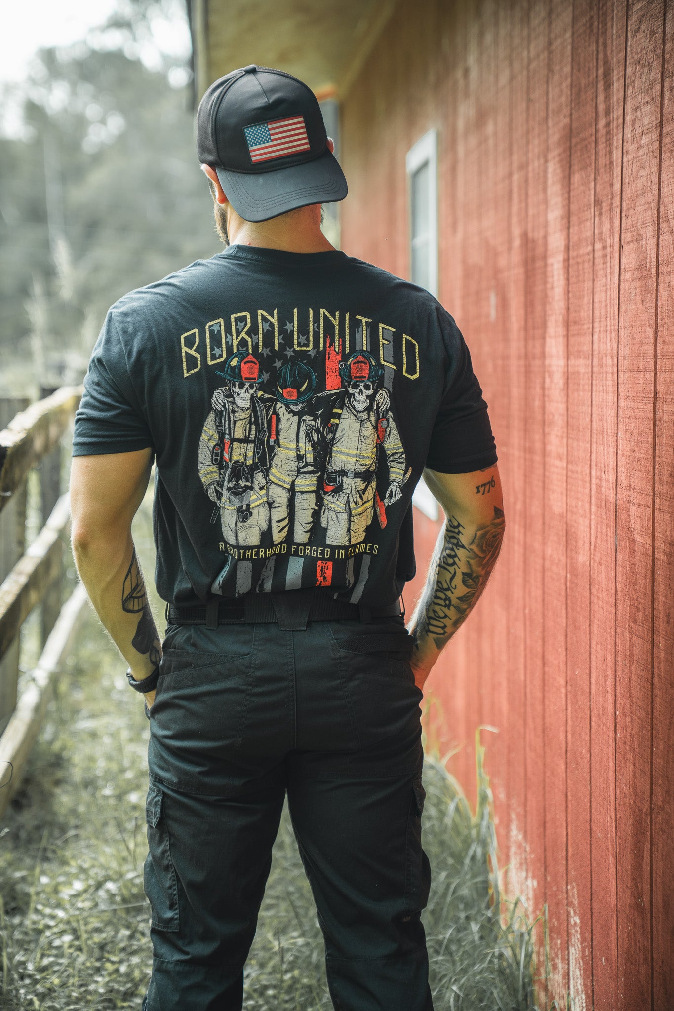 A man wearing a black Born United Private Label T-shirt—Brotherhood Forged In Flames, featuring three masked soldiers on the back—stands outdoors by a red wooden building, facing away from the camera.