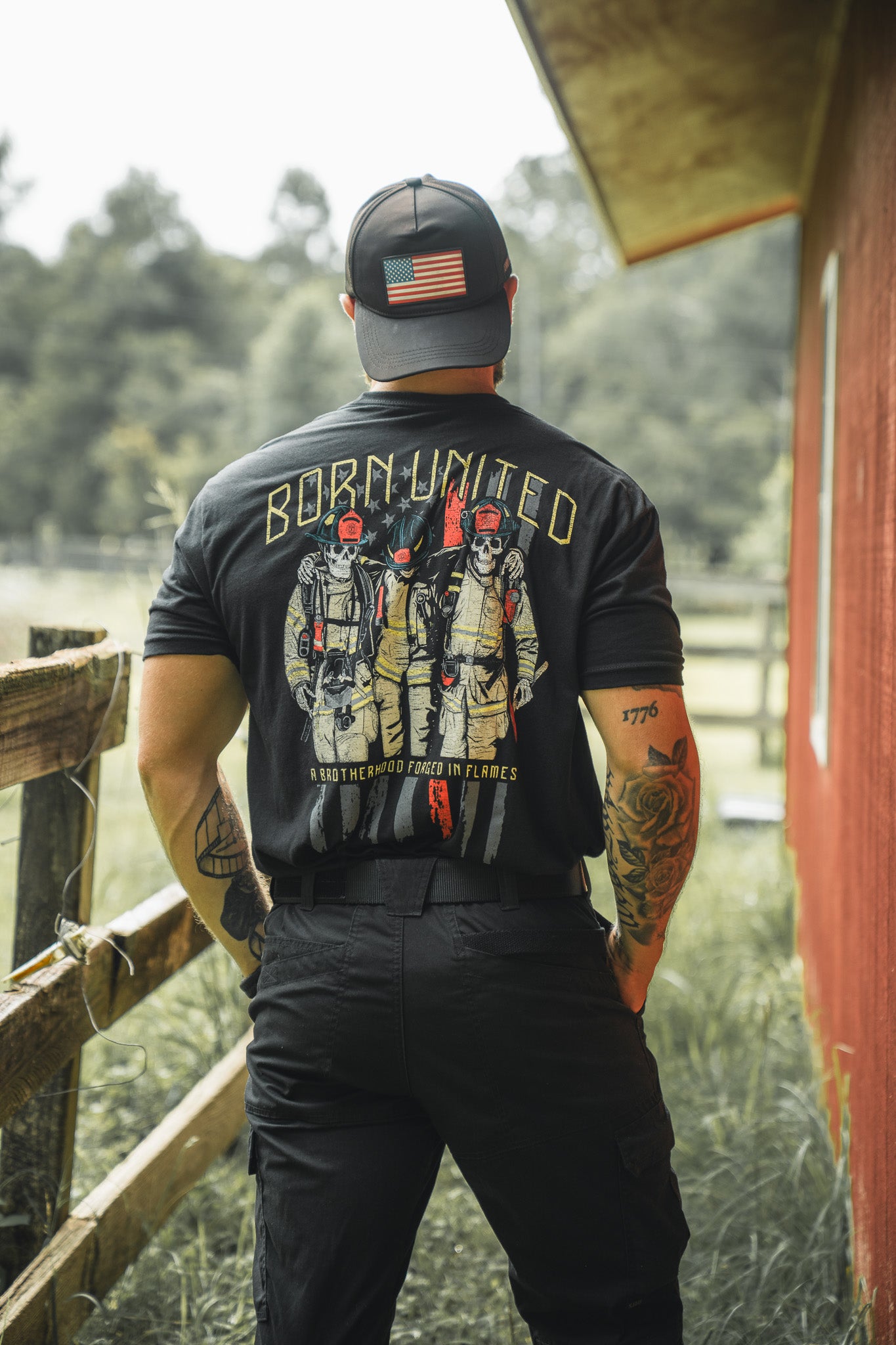A person wearing the Born United Private Label’s Brotherhood Forged In Flames t-shirt with a firefighter graphic stands outdoors, back turned, near a red building and wooden fence. They also wear a black cap with an American flag patch.