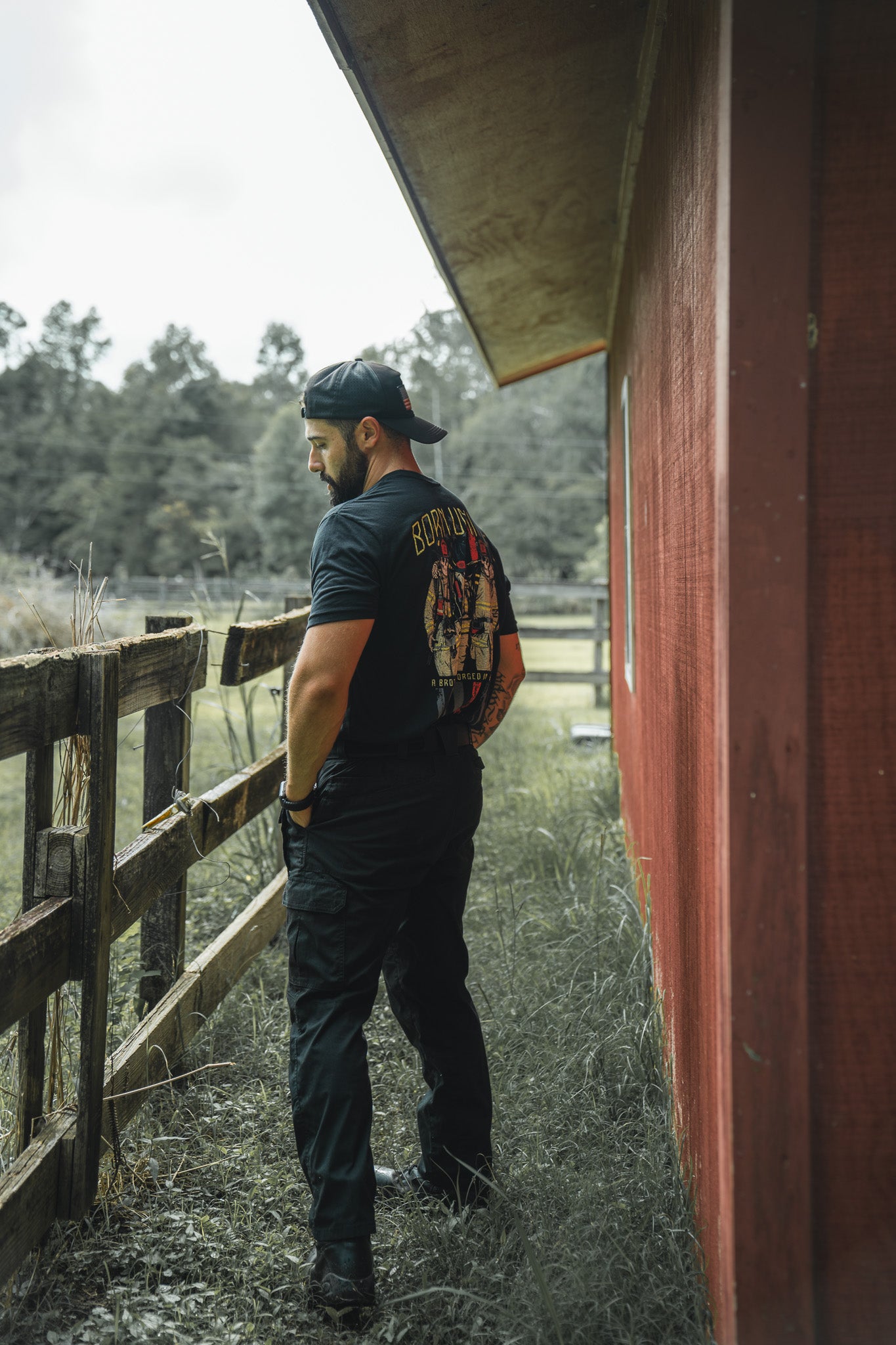 A man in the Brotherhood Forged In Flames black t-shirt by Born United Private Label, black pants, and a backward cap stands by a wooden fence near a red building, looking down with trees behind him.