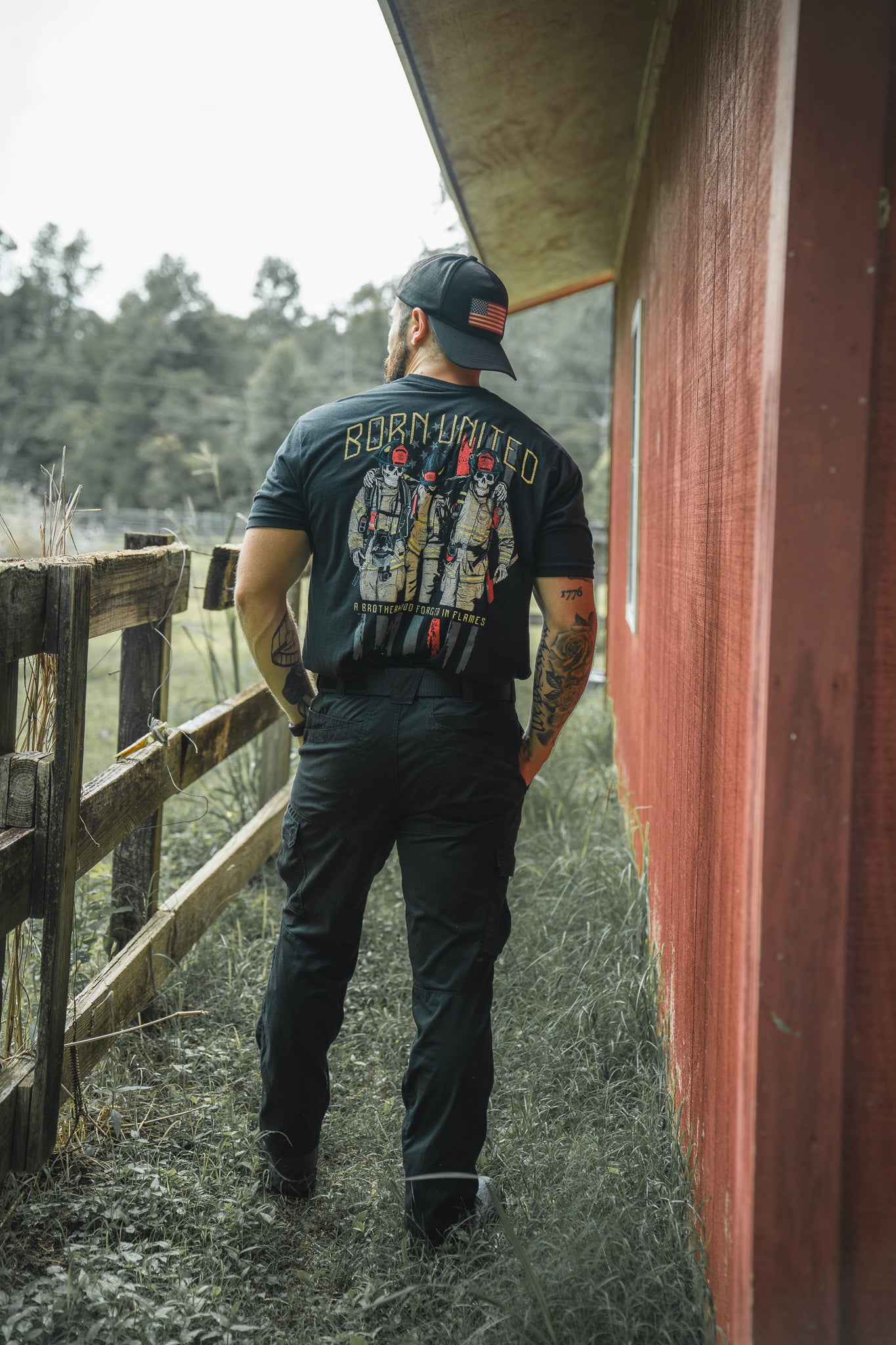 A man wearing a black Brotherhood Forged In Flames tee by Born United Private Label and a black cap walks along a grassy path beside a red wooden building and fence, with trees in the background.