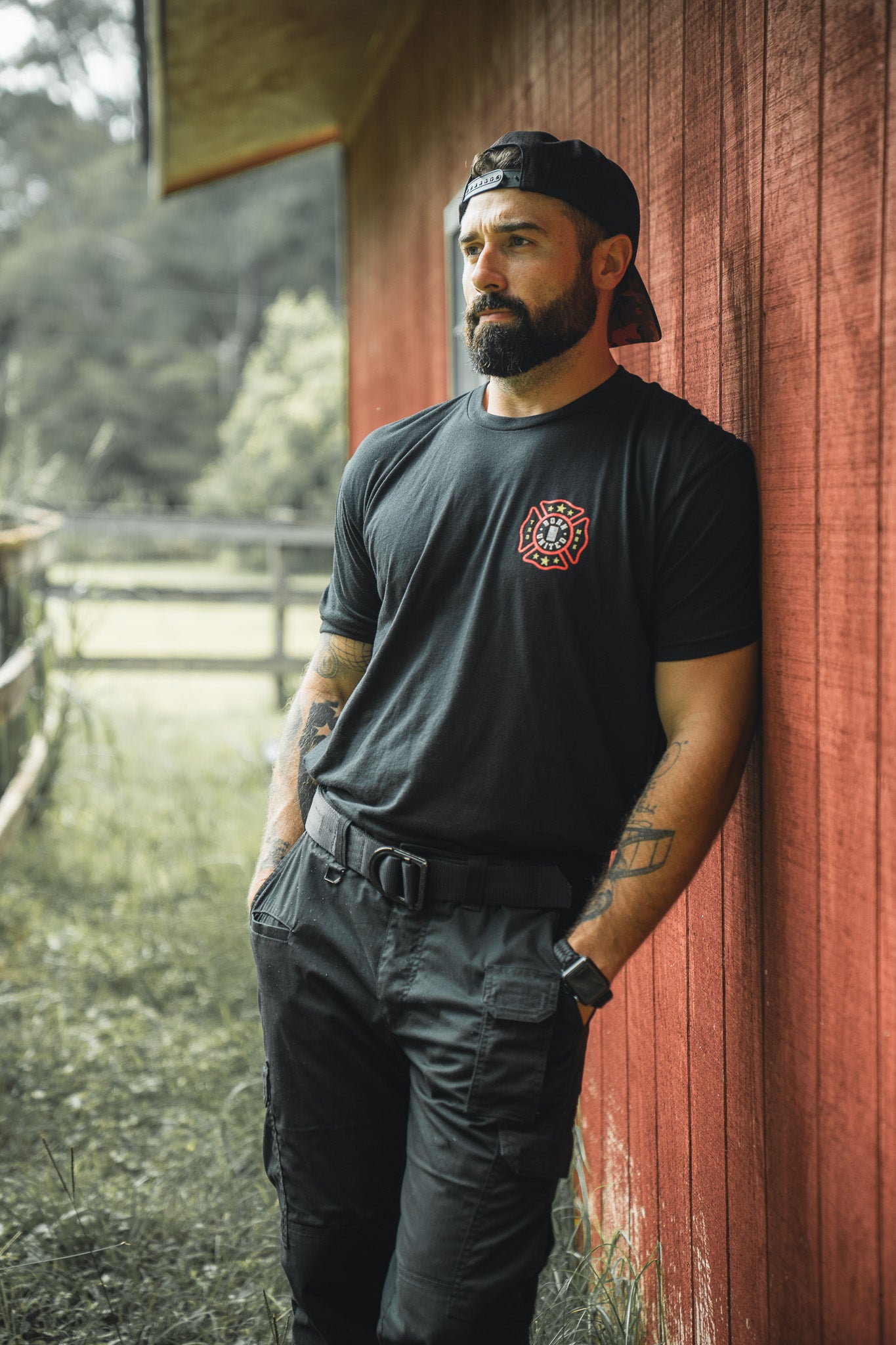 A bearded, tattooed man in a Born United Private Label Brotherhood Forged In Flames black T-shirt, black pants, and a backward cap leans against a red wooden wall outdoors, gazing thoughtfully into the distance.
