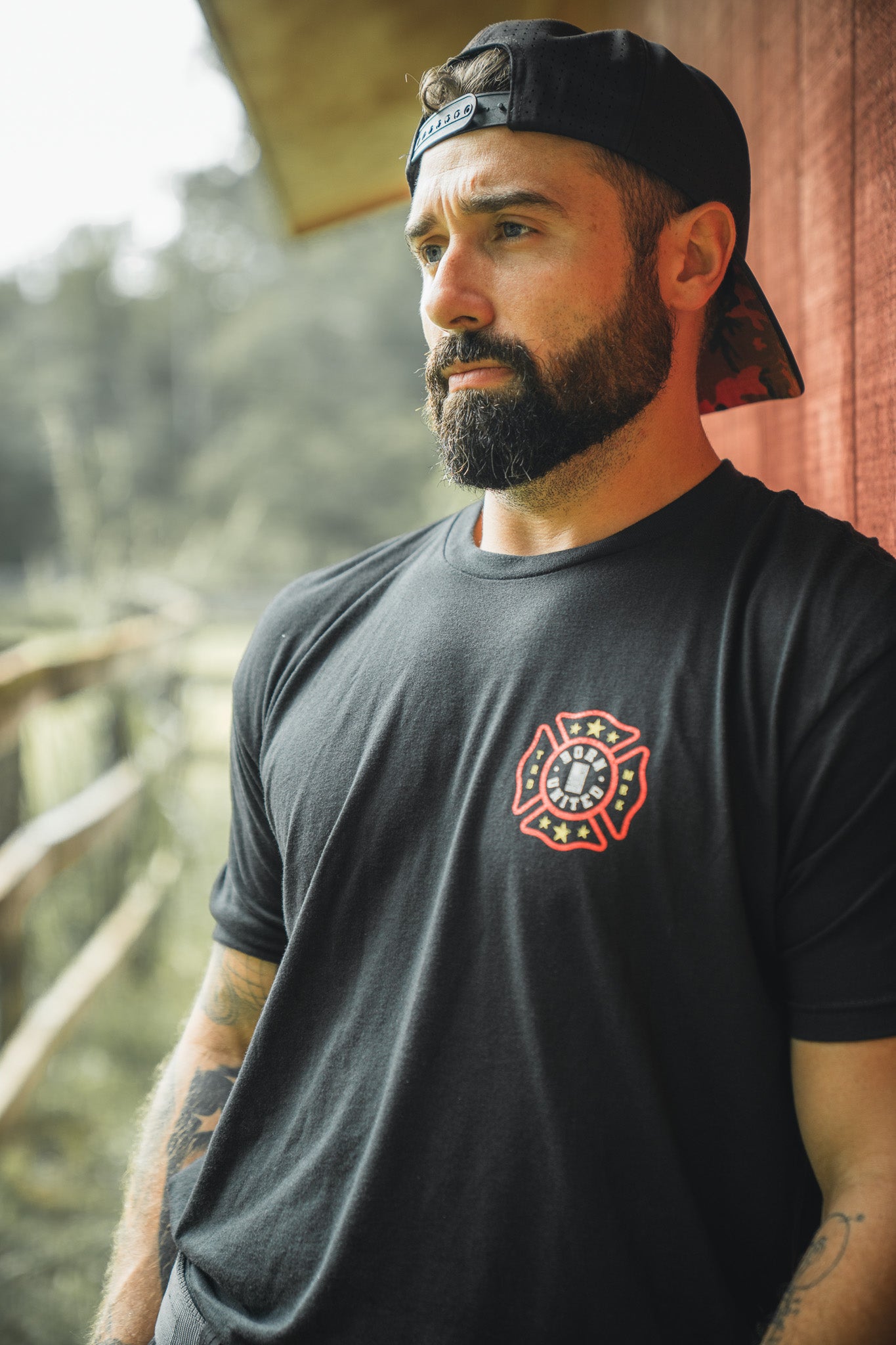 A bearded man in a backward black cap and Born United Private Labels Brotherhood Forged In Flames T-shirt stands outdoors by a wooden fence and red building, gazing thoughtfully into the distance.