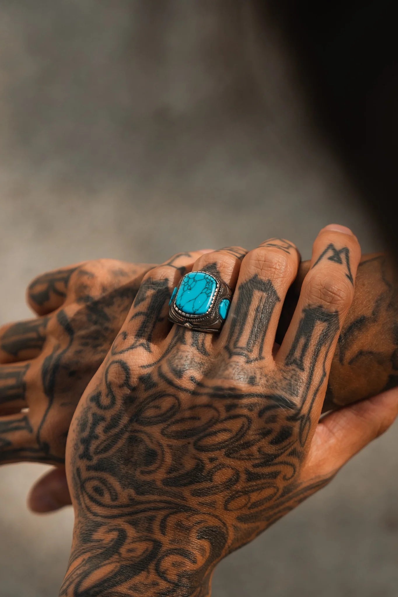 A tattooed hand wearing the Desperado Ring by Relentless Betrayal, featuring a turquoise stone in detailed silver, gently holds another tattooed hand. Both are adorned with handcrafted jewelry. The serene scene is complemented by a blurred neutral background.