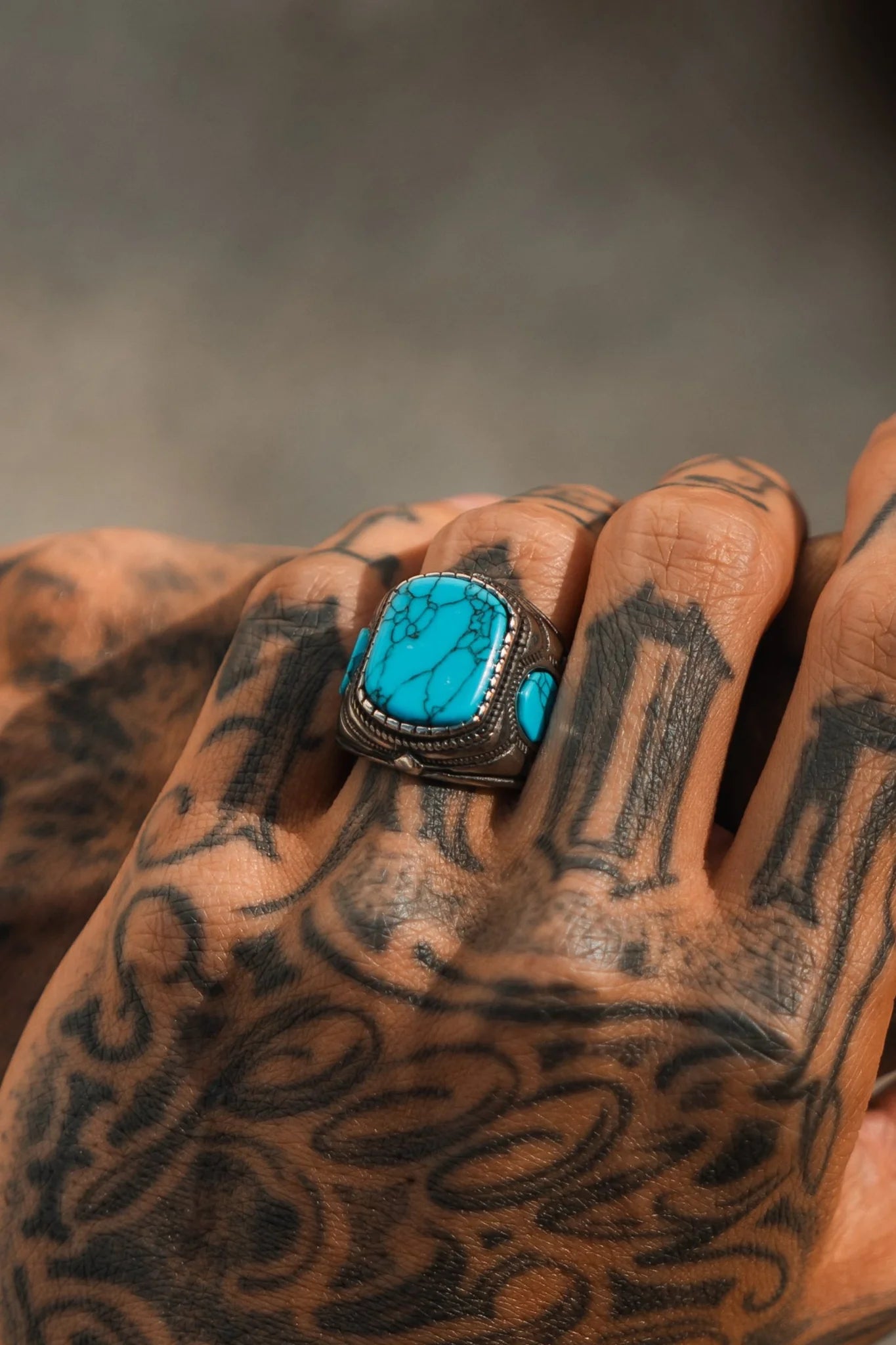 A tattooed hand showcases the Desperado Ring by Relentless Betrayal, featuring a bright turquoise stone with intricate natural vein patterns. The background is blurred, enhancing the rings allure amid intricately tattooed fingers and hand.