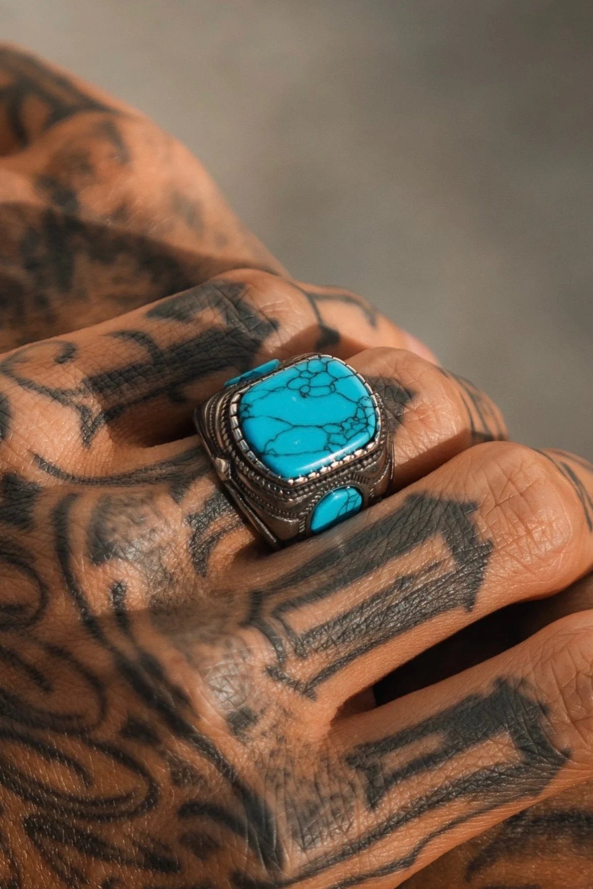 A close-up shows a tattooed hand adorned with Relentless Betrayals Desperado Ring, featuring intricate stainless steel craftsmanship paired with a vibrant turquoise stone.