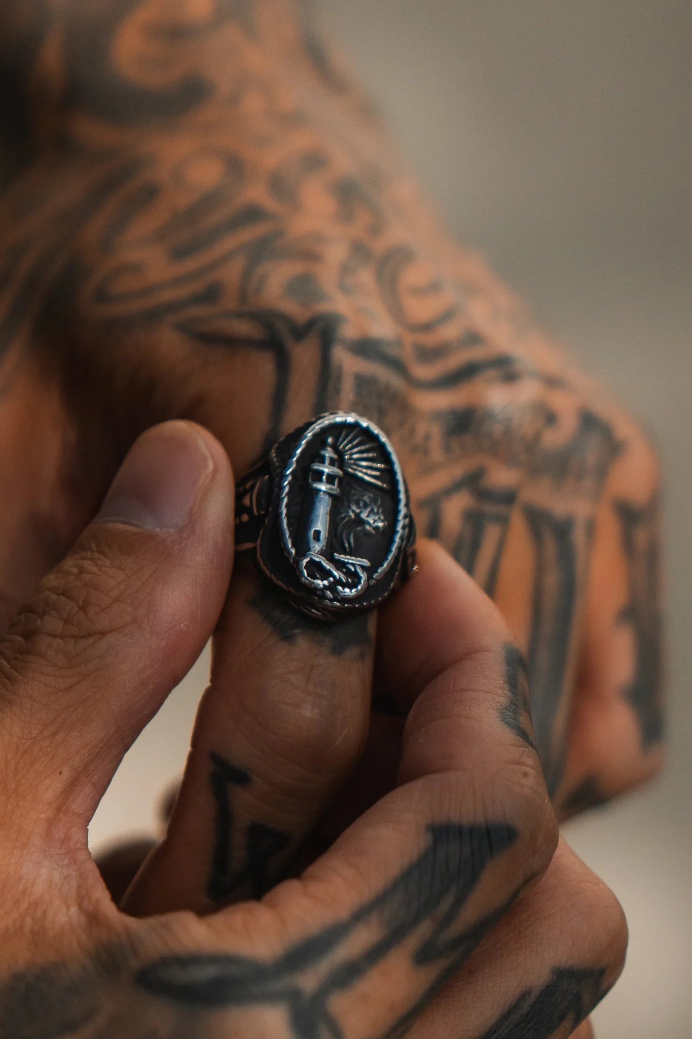 A tattooed hand holds the Guide My Reckless Soul Ring by Relentless Betrayal, with its intricate lighthouse designs captivating attention against a blurred background.