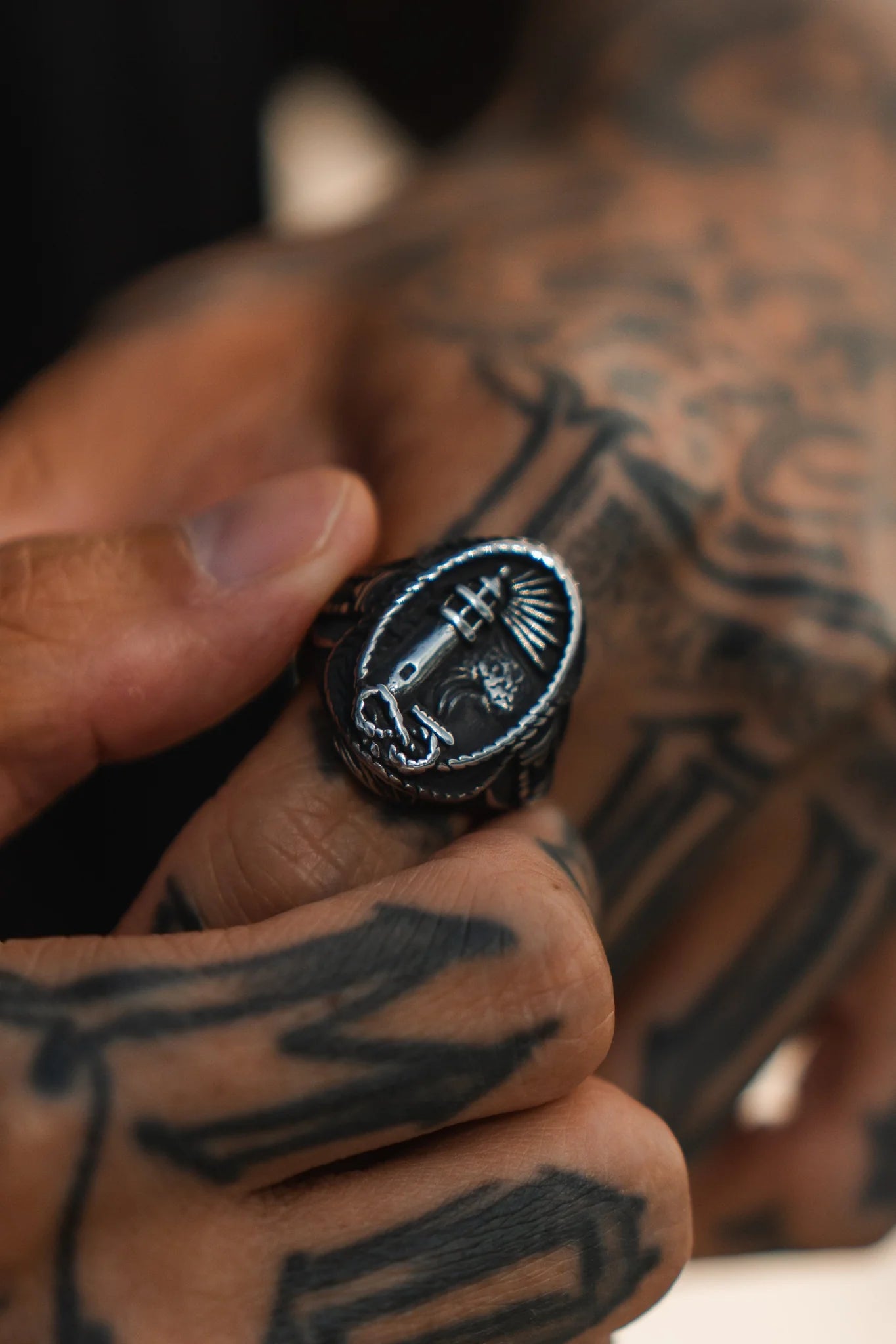 A tattooed hand holds a handcrafted Guide My Reckless Soul Ring by Relentless Betrayal, featuring engravings of a whale and ships wheel, with fingers displaying intricate tattoos.