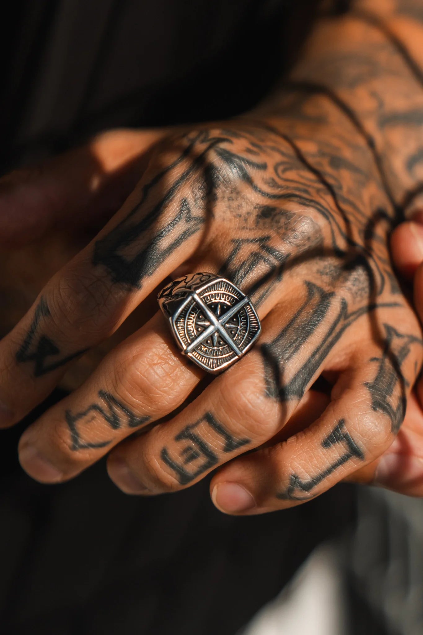 A tattooed hand displays the handcrafted True North Ring by Relentless Betrayal, a square-shaped stainless steel piece on the middle finger with a detailed pattern and central cross design. The background is slightly blurred to highlight the hand and ring.