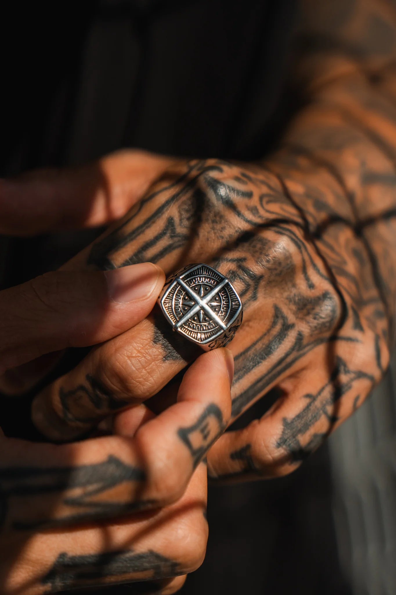 A person with tattooed hands holds the True North Ring by Relentless Betrayal, showcasing a black, geometric design set in a silver border. Made from stainless steel, the rings intricate details catch dramatic shadows and light.