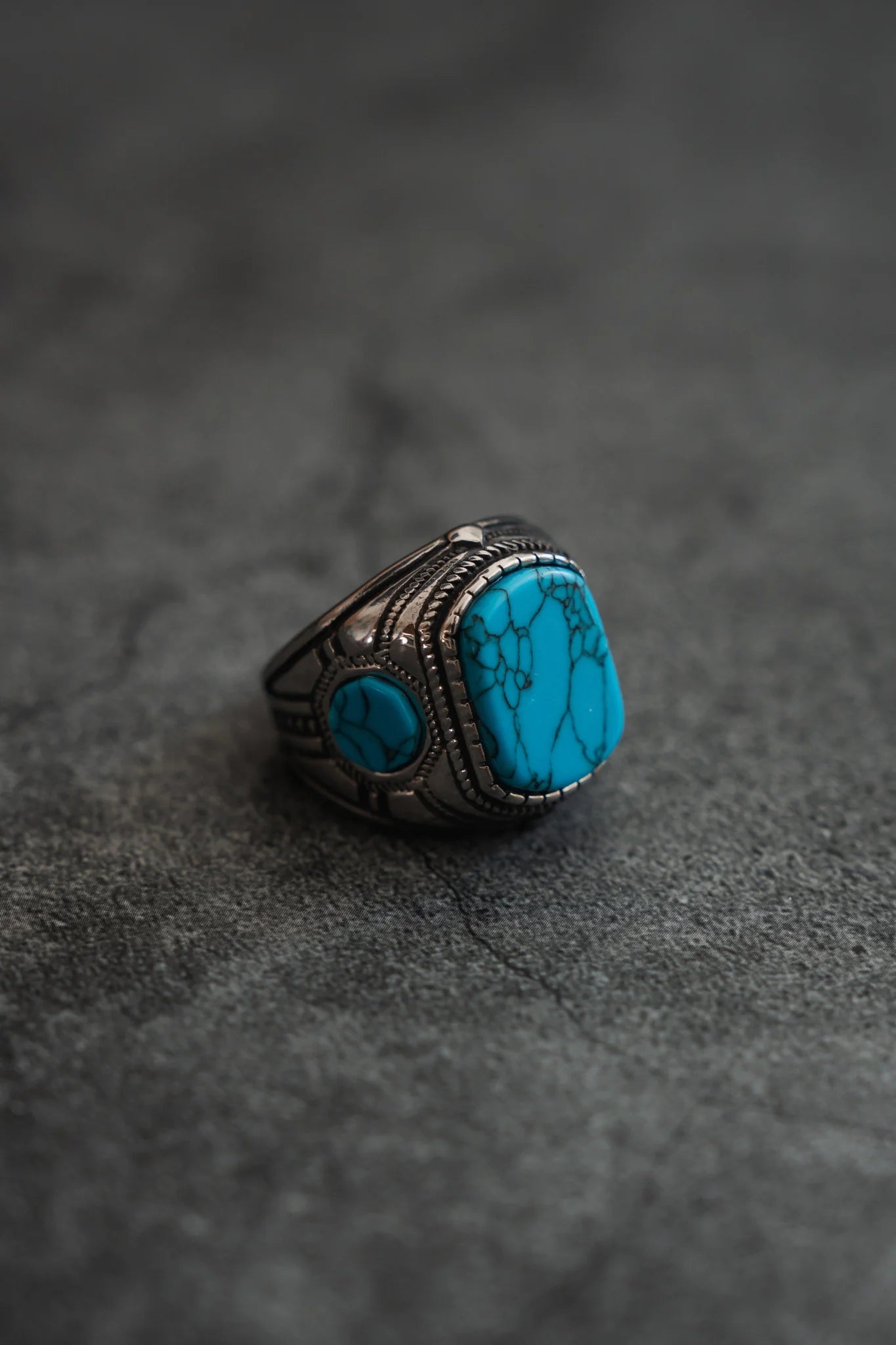 The Desperado Ring by Relentless Betrayal is a handcrafted masterpiece, featuring a large square turquoise stone with distinct veins set against intricate silver detailing on a textured gray surface.