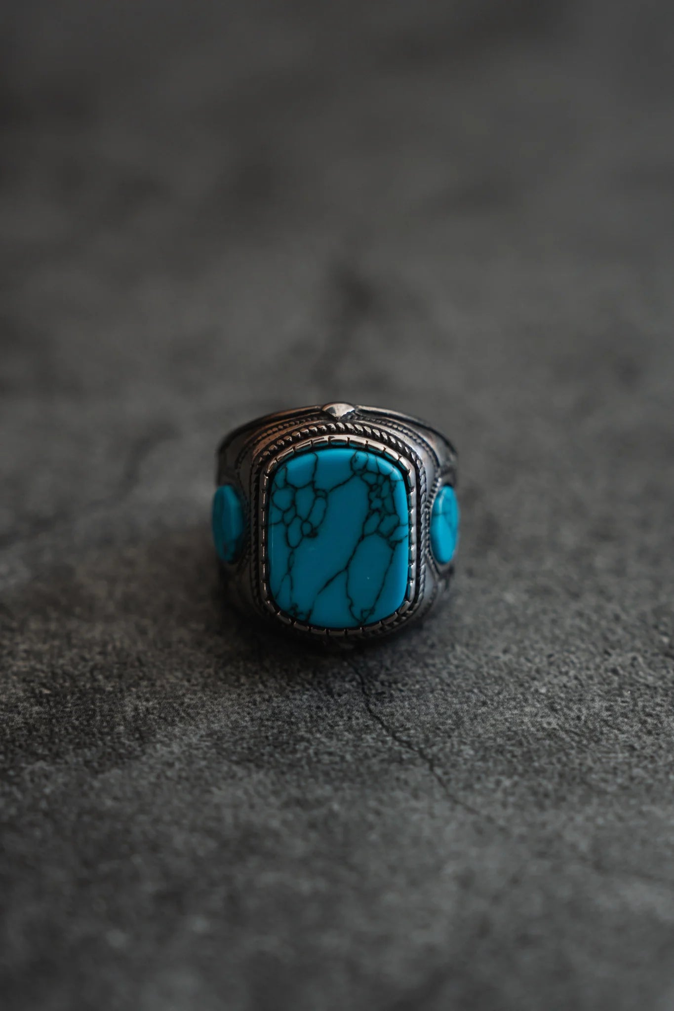 The Desperado Ring from Relentless Betrayal is a handcrafted silver piece featuring a large rectangular turquoise stone with intricate veining, set in a textured band and elegantly displayed on a gray textured surface.