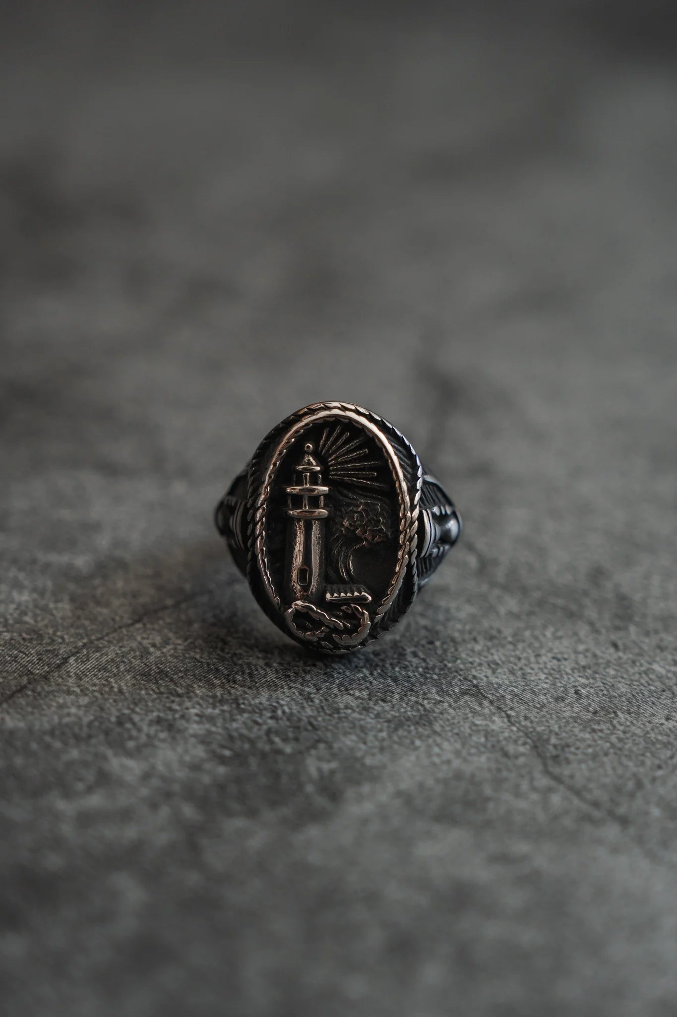 The Guide My Reckless Soul Ring by Relentless Betrayal features a handcrafted silver design with an engraved lighthouse and upright snake, elegantly displayed on a textured gray surface.