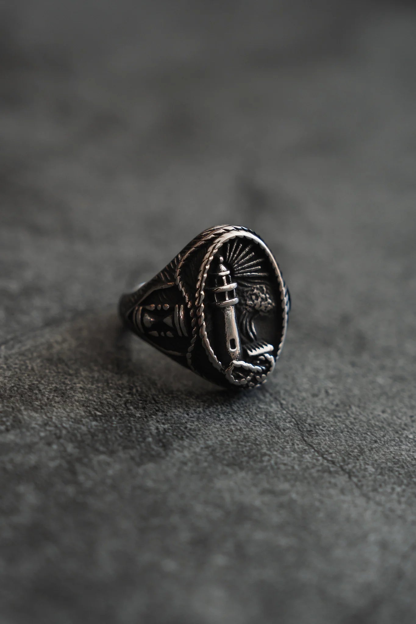 The Guide My Reckless Soul Ring by Relentless Betrayal is a handcrafted silver piece with a lighthouse engraved in an oval frame, adorned with ornate rope patterns against a dark, textured backdrop—ideal for those seeking unique elegance.
