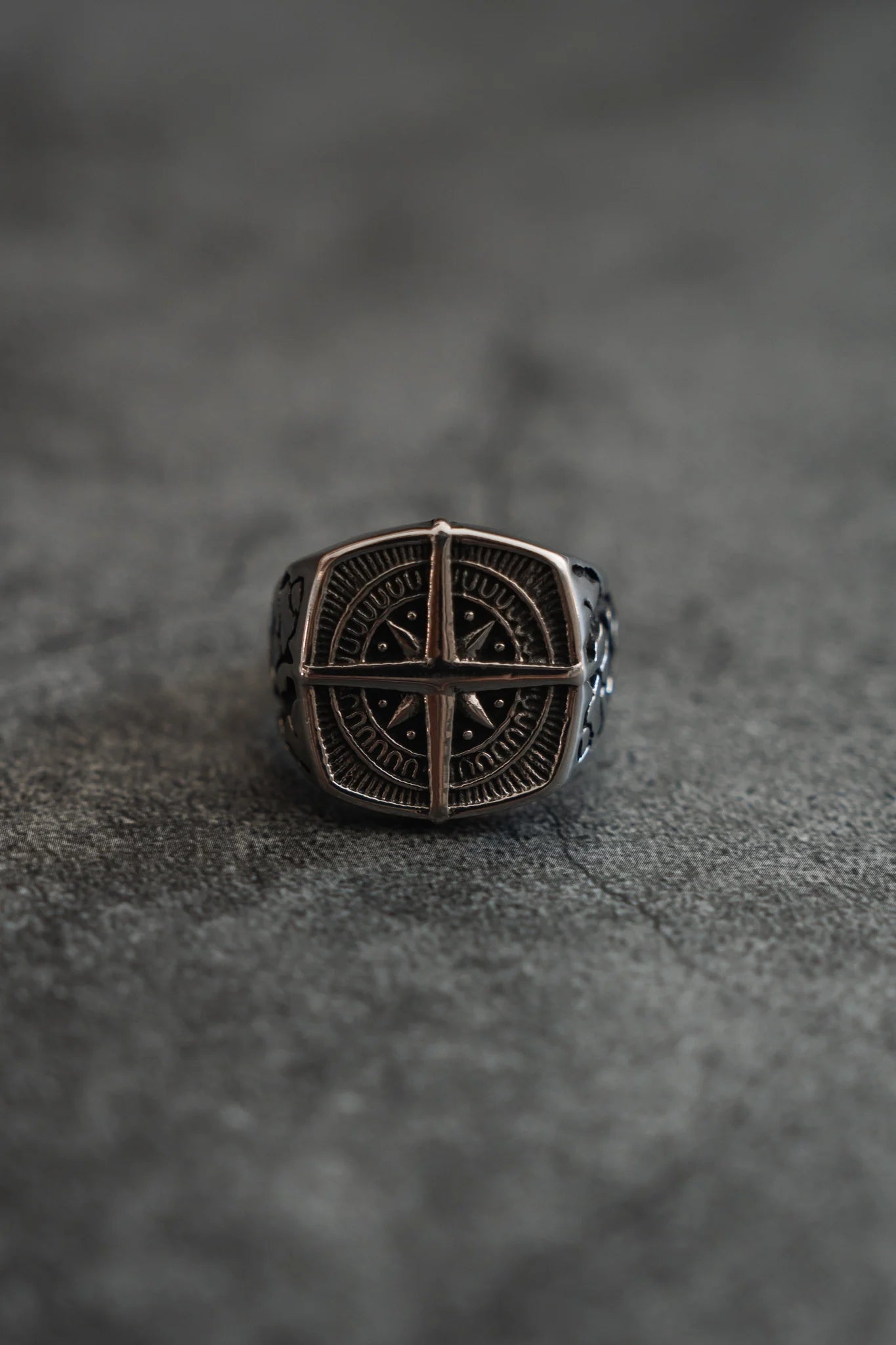 The Relentless Betrayal True North Ring showcases a handcrafted silver design with an intricate circular pattern and a cross-like symbol at its center, all set against a smooth dark gray surface.