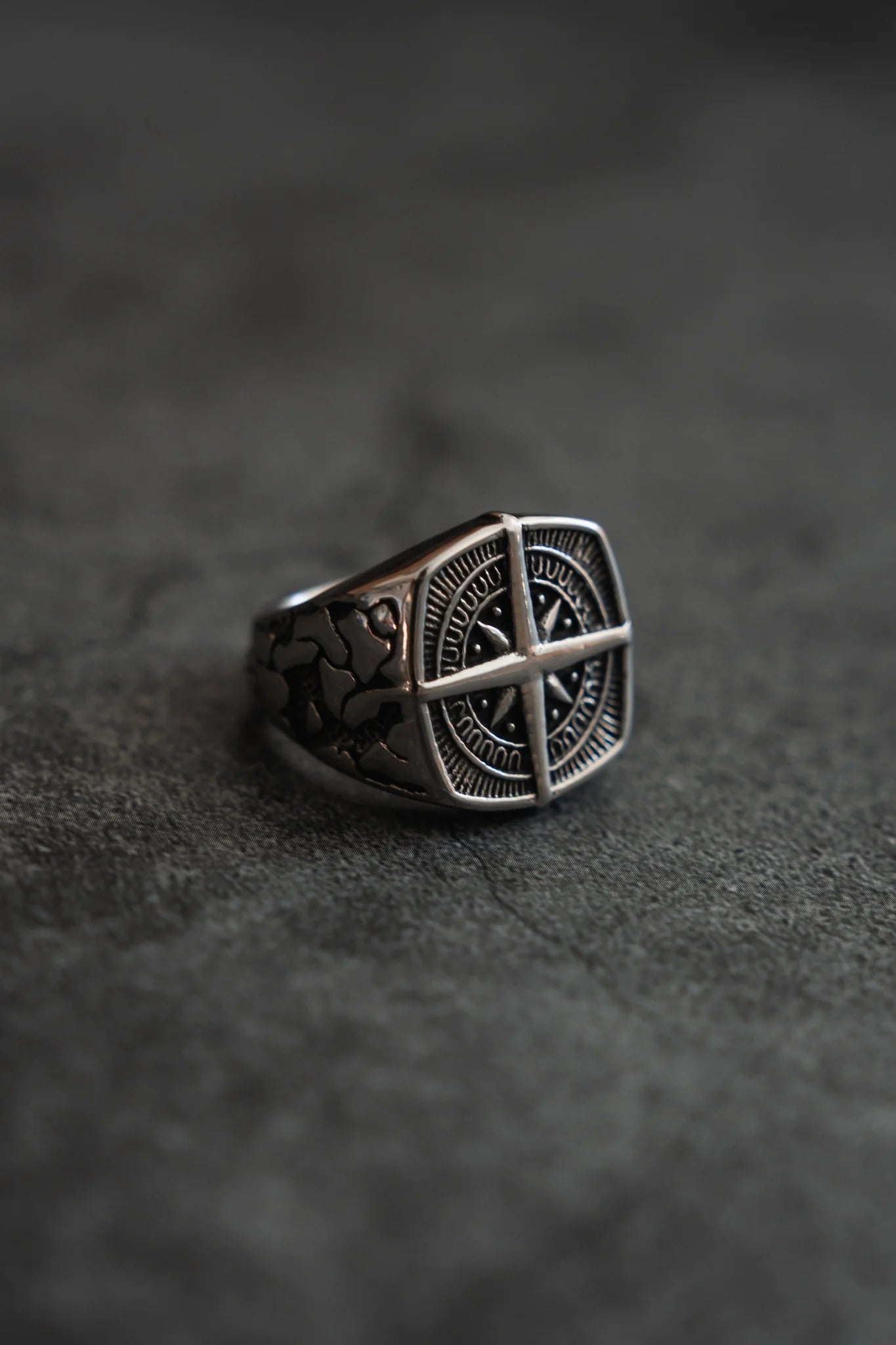 The True North Ring by Relentless Betrayal is a stunning stainless steel piece, featuring an ornate circular emblem with a cross-like design. Its band displays intricate patterns on a textured gray background.