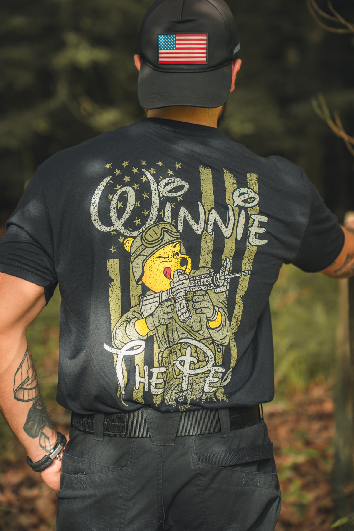 A patriot in a black cap with an American flag patch and a T-shirt from Born United Private Label features the Winnie the Pew design: a storybook bear in military gear holding a rifle, with Winnie The Pew printed on the back.