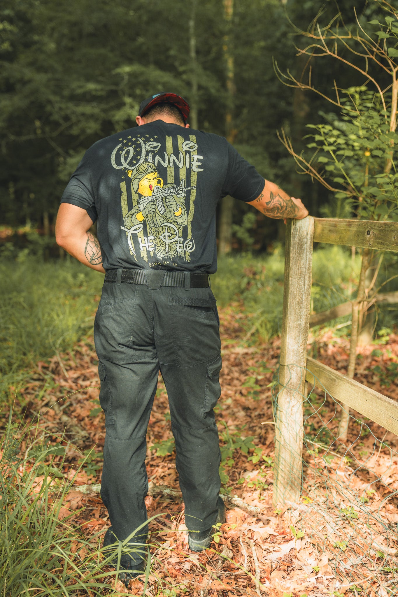 A person stands on a sunlit forest path, wearing the black Winnie the Pew t-shirt by Born United Private Label, hand resting on a wooden fence, capturing the storybook bears adventurous spirit.