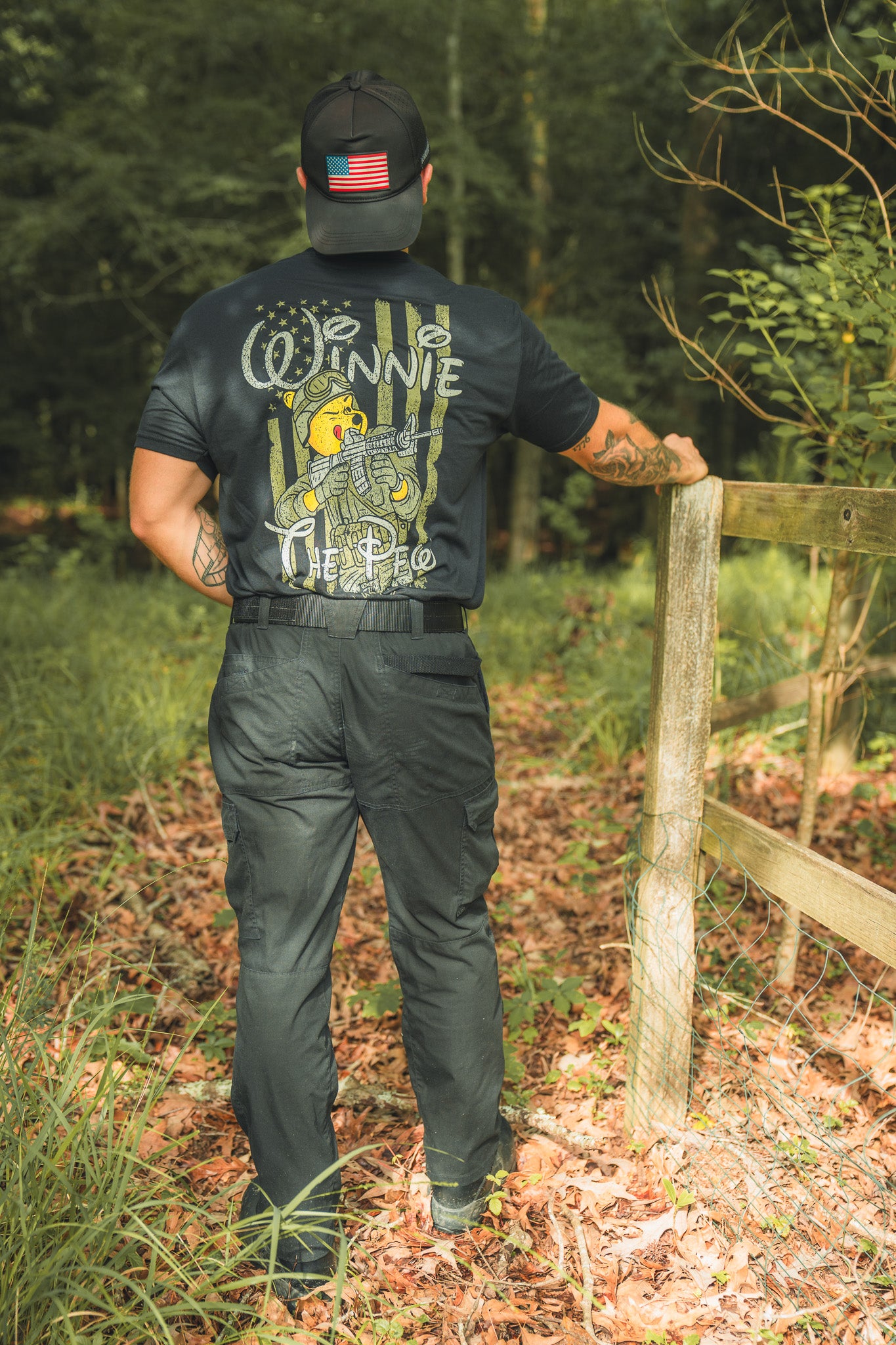 Standing outdoors by a wooden fence, this patriot wears a black Winnie the Pew shirt from Born United Private Label with a storybook bear graphic and a cap featuring an American flag patch, surrounded by trees and fallen leaves.