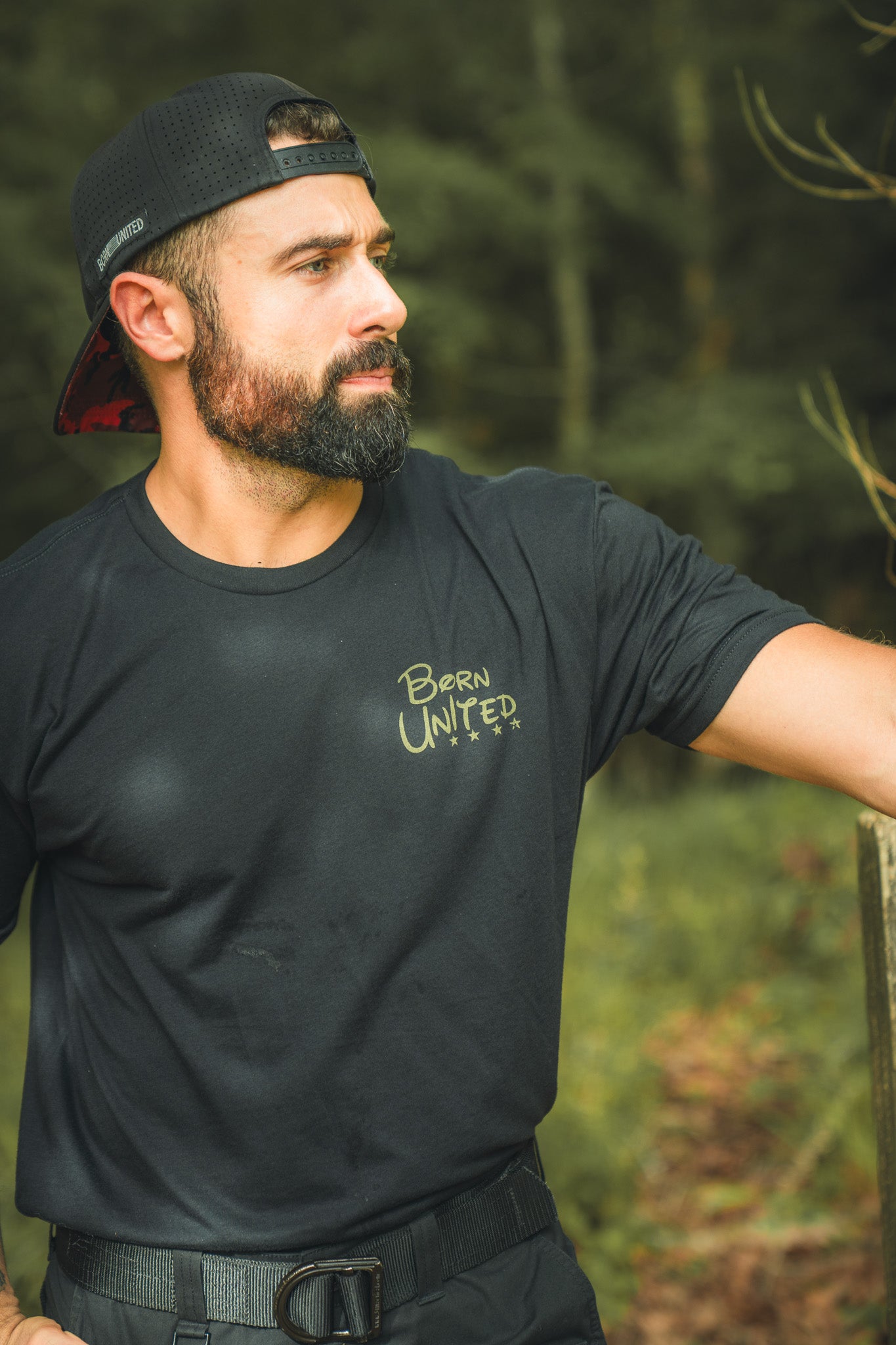 A bearded man in a black cap and a Winnie the Pew Born United Private Label t-shirt stands outdoors by a wooden fence, gazing thoughtfully aside with a blurred forest background.