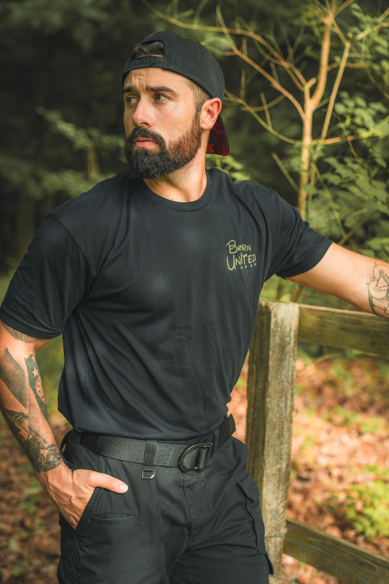 A bearded man in a black Winnie the Pew T-shirt by Born United Private Label and cap stands by a wooden fence outdoors, dressed in black pants with tattooed arms, surrounded by greenery and trees.