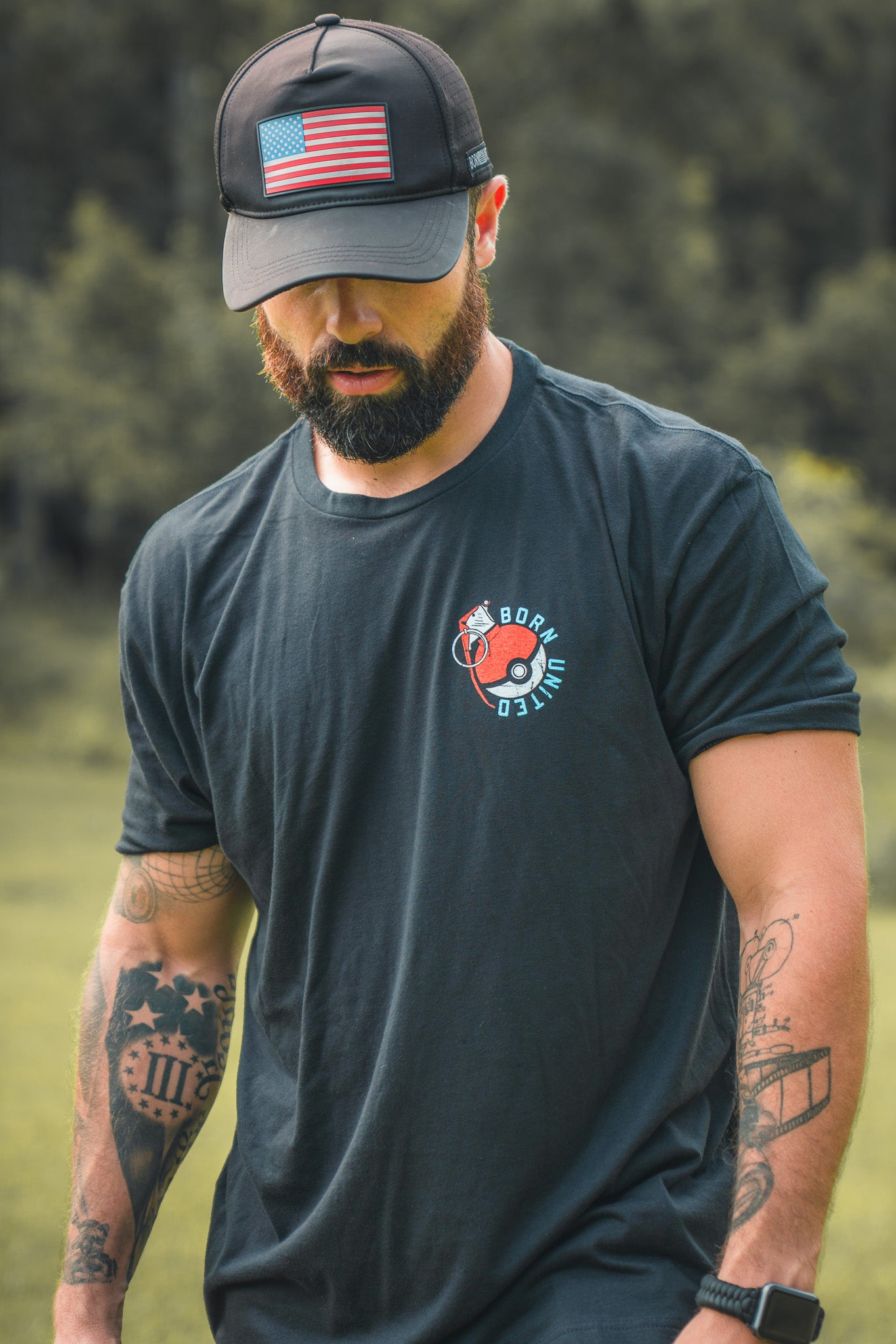A bearded man in a black cap with an American flag patch and a Born United Private Label “Pika-Pew” tee stands outdoors, tattooed arms visible, looking down as if searching for collectibles. The background shows blurred greenery.