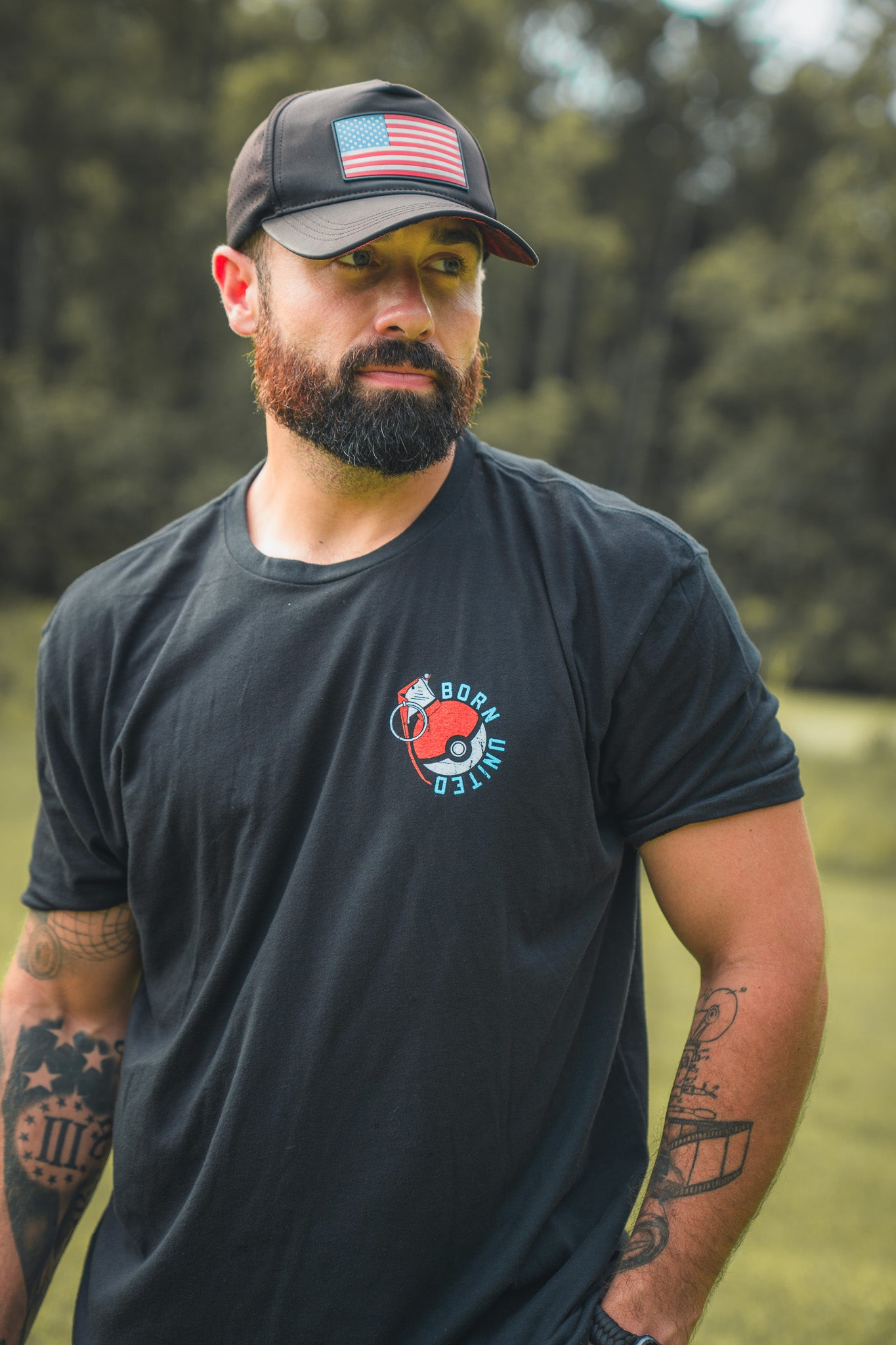 A bearded man with tattooed arms wears the Born United Private Label Pika-Pew black T-shirt and an American flag cap, standing outside in a green, blurred setting, looking to the side—ready for adventure.