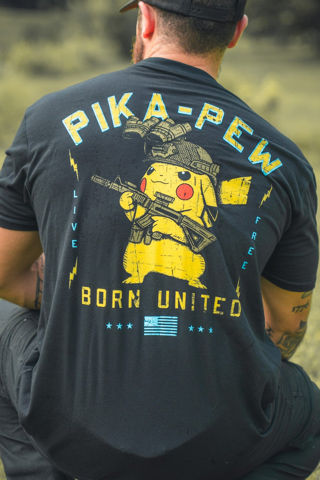A man kneels on grass, facing away, wearing a black Born United Private Label Pika-Pew T-shirt with a Pikachu holding a rifle and bold PIKA-PEW, BORN UNITED, and LIVE FREE text—perfect for Pokémon collectors.