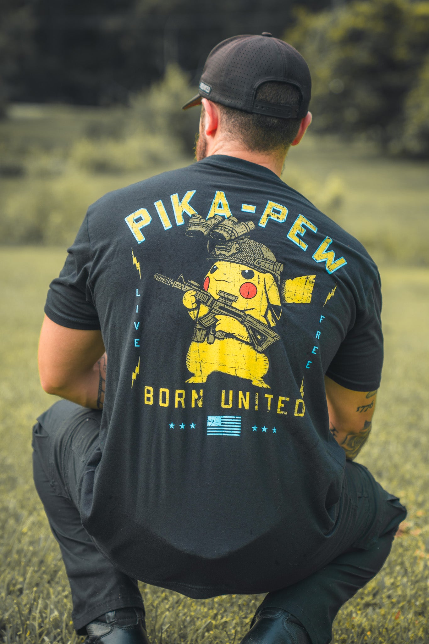 A man kneels on grass, facing away, wearing a black Born United Private Label Pika-Pew T-shirt with a Pikachu holding a rifle and bold PIKA-PEW, BORN UNITED, and LIVE FREE text—perfect for Pokémon collectors.